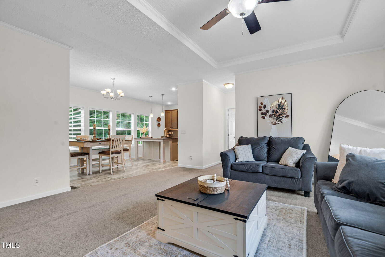 Open living area with gray sofas, a large mirror, a coffee table, and framed wall art; dining table and kitchen with wooden cabinets visible in the background; ceiling fan overhead, neutral colors throughout.