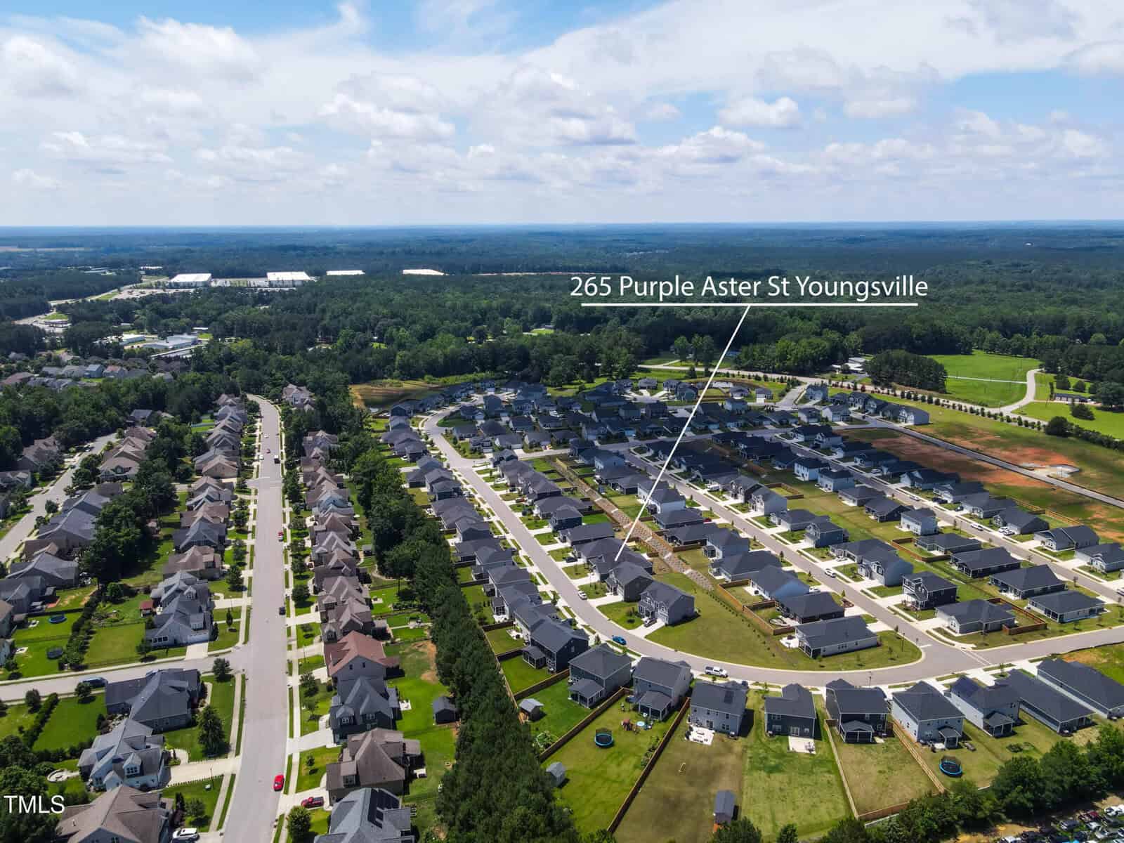 Aerial view of a suburban neighborhood with rows of houses and green lawns. A label with a line points to one specific house, identified as 265 Purple Aster St, Youngsville. The area is surrounded by trees and open land.