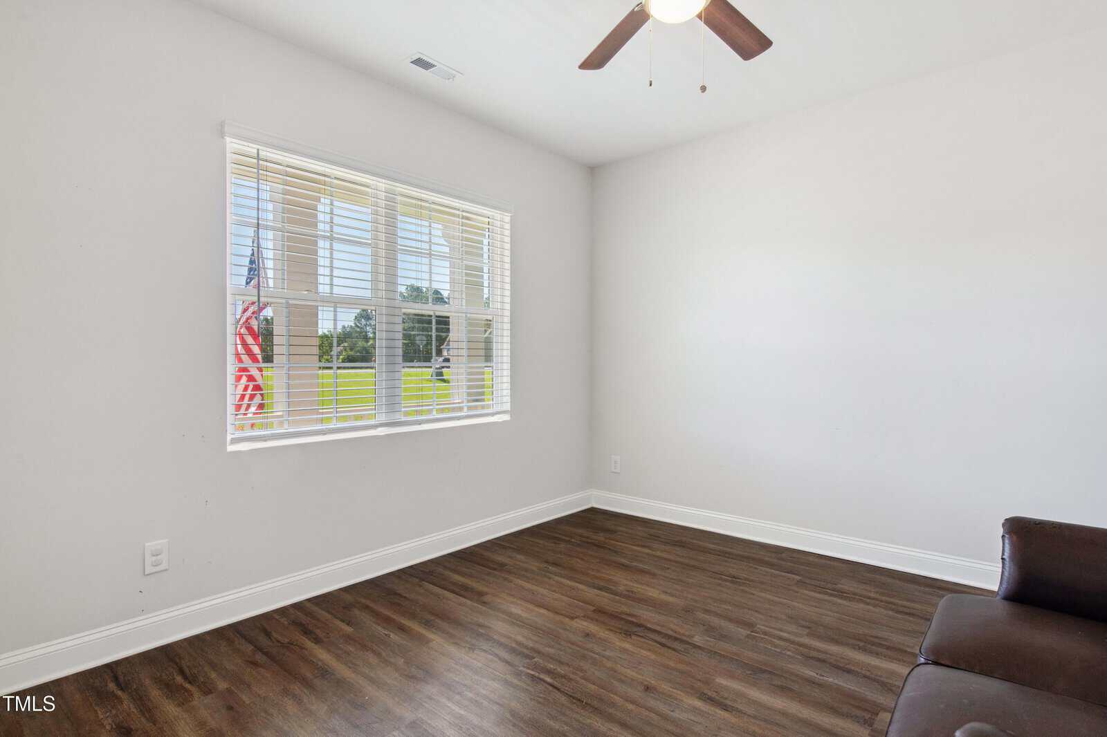 A mostly empty room with white walls, a large window with blinds, wood flooring, a ceiling fan with a light, and a small part of a brown couch visible in the lower right corner. An American flag is seen outside the window.