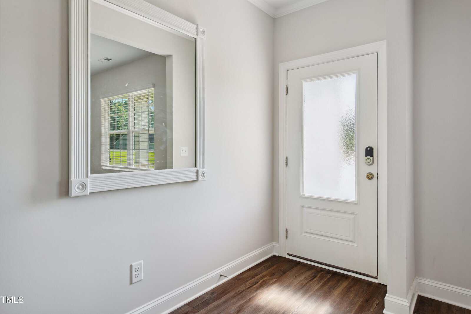 A white front door with a frosted glass panel is next to a large rectangular wall mirror with a white frame. The floor is dark wood, and the walls are painted light gray. Sunlight enters through a window reflection.