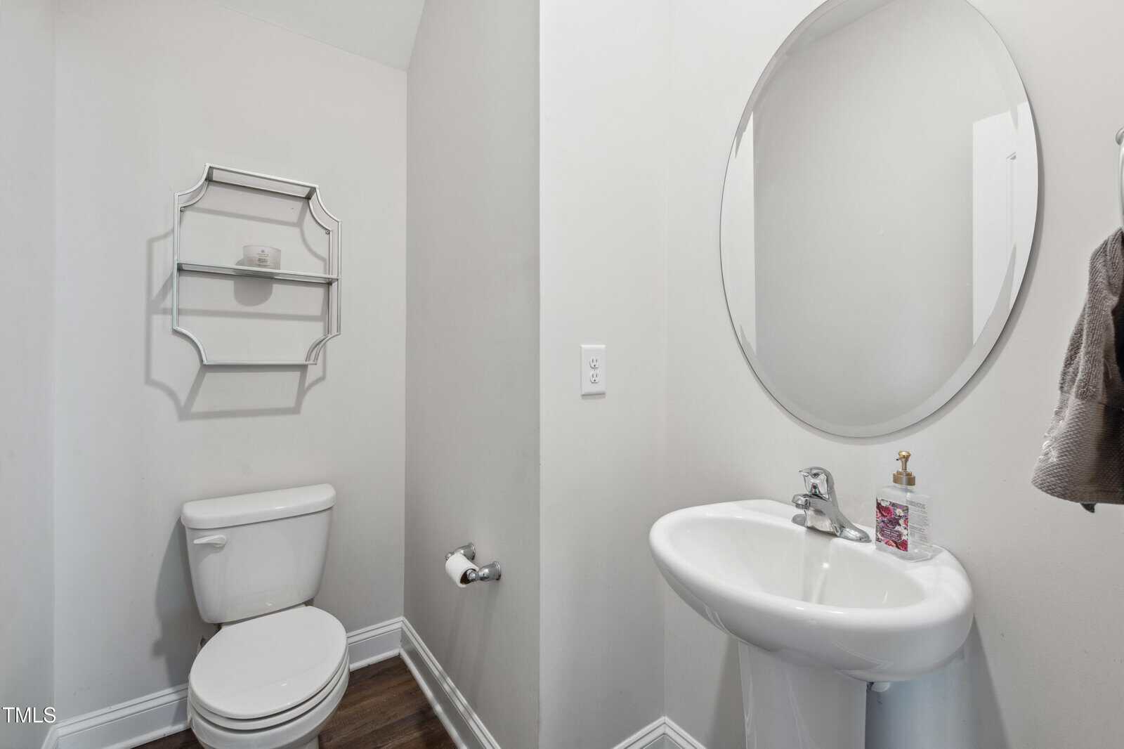 A bathroom with a white toilet, a wall-mounted metal shelf, a white pedestal sink with a round mirror above it, a towel hanging on the wall, and a soap dispenser on the sink. The floor is dark wood.