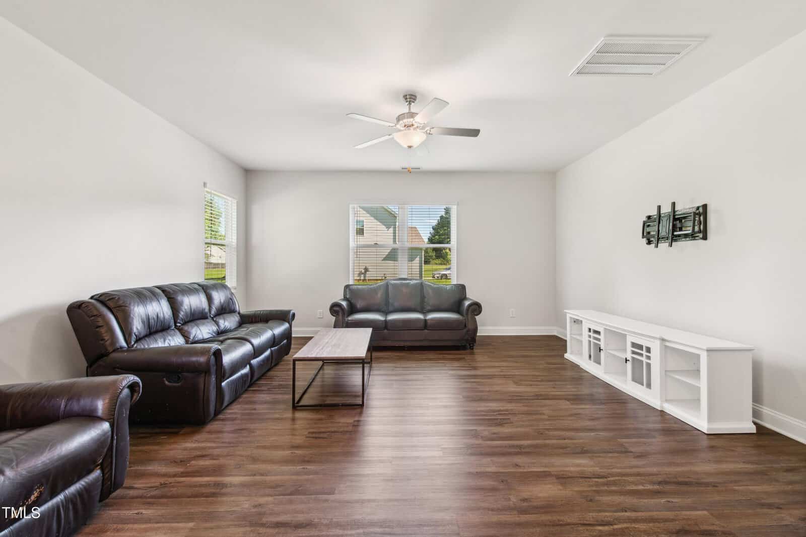 A living room with dark brown leather sofas, a wooden coffee table, white walls, large windows, a ceiling fan, a white TV console, and a wall-mounted TV bracket on wood flooring.