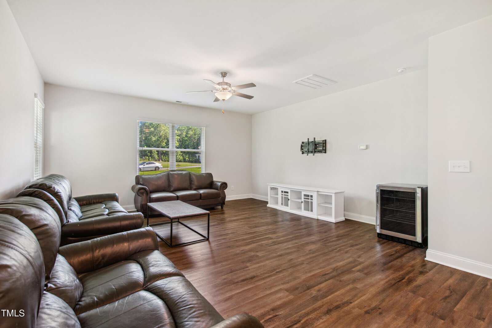 Spacious living room with brown leather sofas, a coffee table, a ceiling fan, large window with blinds, white TV stand, wall-mounted TV, and dark wood flooring.