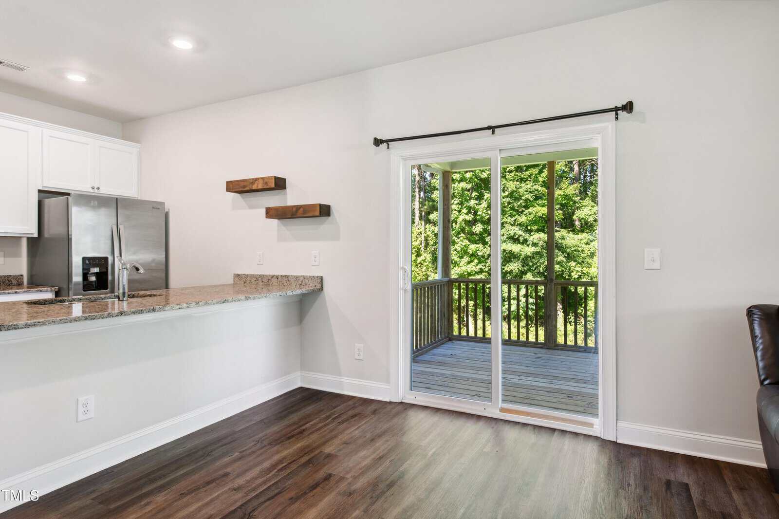 A bright kitchen with white cabinets, stainless steel refrigerator, granite countertop, and wooden shelves. Sliding glass doors open to a wooden deck with a view of green trees outside.