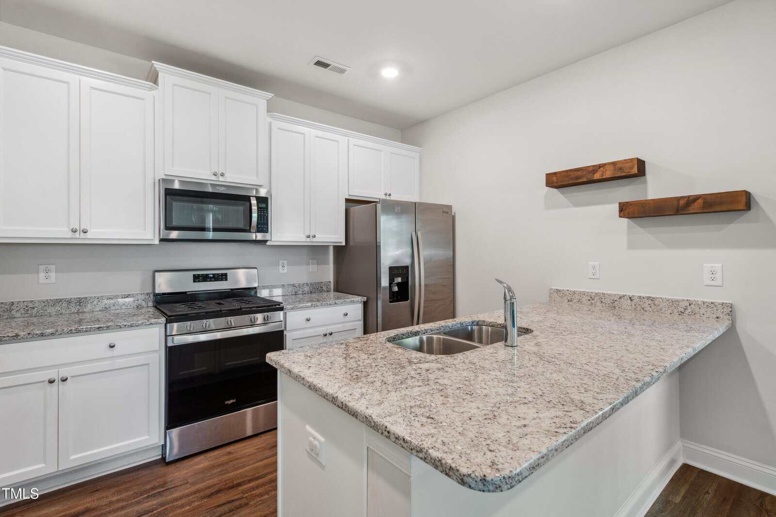 Modern kitchen with white cabinets, stainless steel appliances, granite countertops, a built-in microwave above the stove, a double-door refrigerator, a sink on a central island, and two wooden wall shelves.