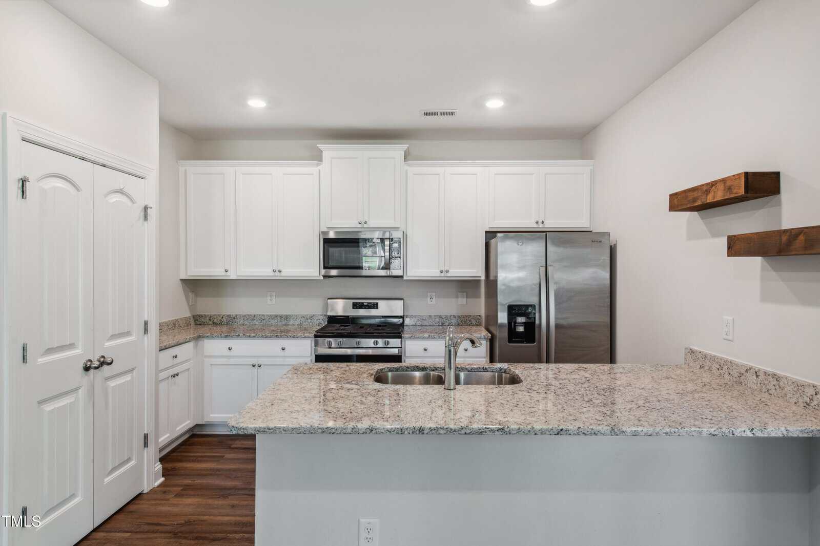 A modern kitchen with white cabinets, stainless steel appliances, a double sink, granite countertops, wooden floating shelves on the right wall, and wooden floors. The space is brightly lit with ceiling lights.