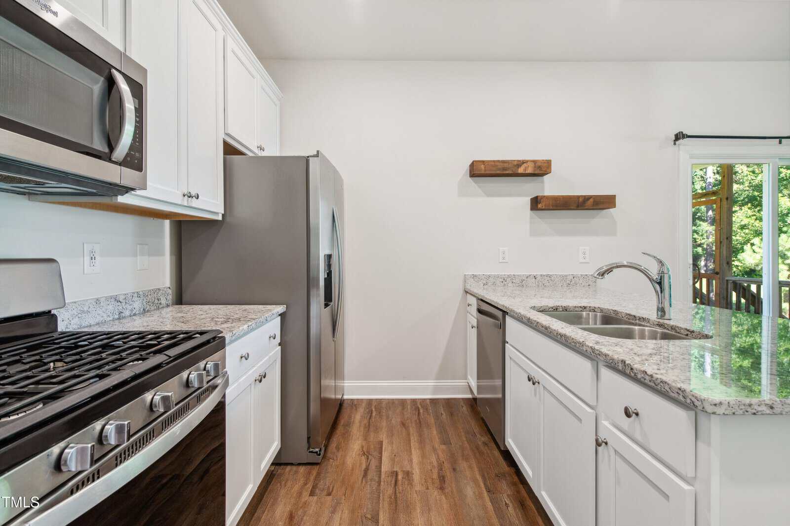 A modern kitchen with white cabinets, stainless steel appliances, granite countertops, a gas stove, wood floors, two wooden floating shelves, and a large window overlooking a green outdoor area.