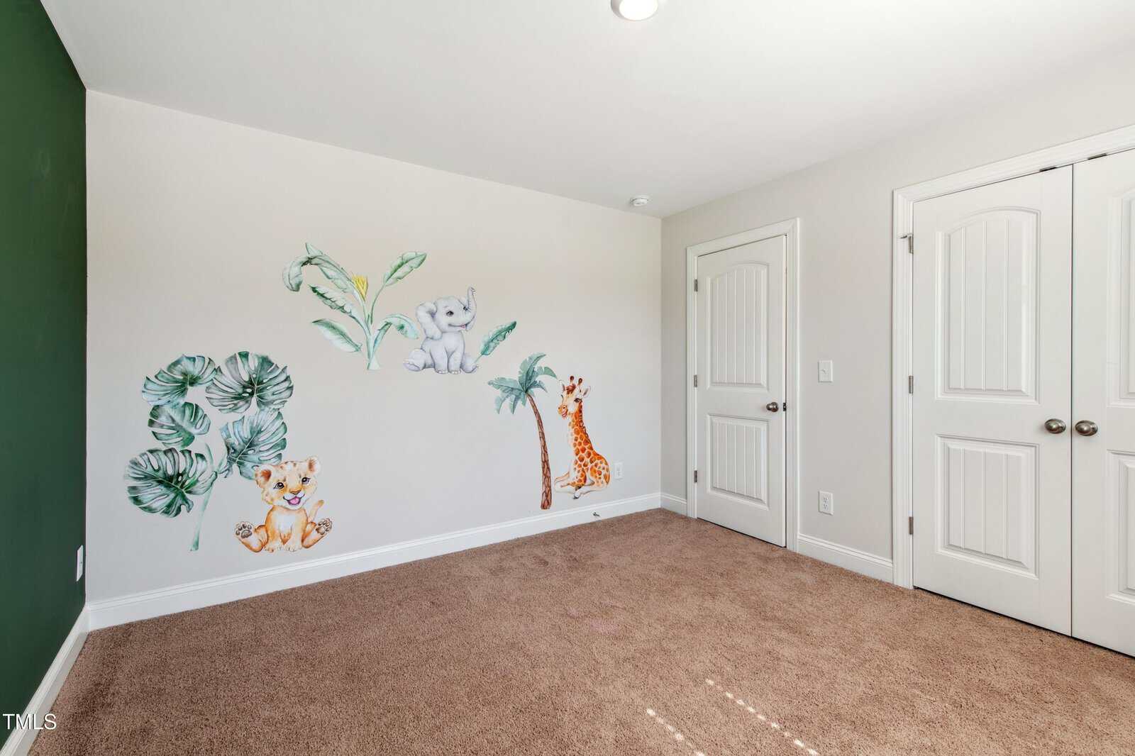 Empty room with beige carpet, green accent wall, and a mural on another wall featuring painted animals—an elephant, tiger, and giraffe—alongside tropical plants and trees. Double white doors are in the corner.