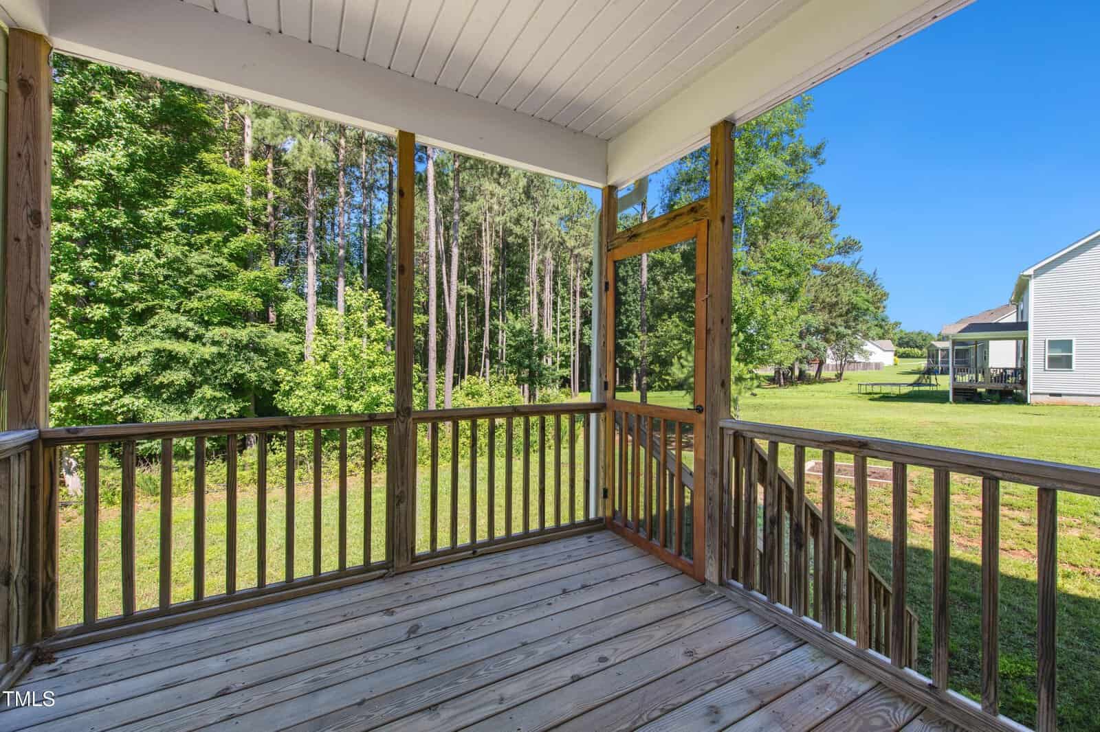 A covered wooden porch with railings overlooks a grassy backyard and trees. Neighboring houses are visible in the background under a clear blue sky.