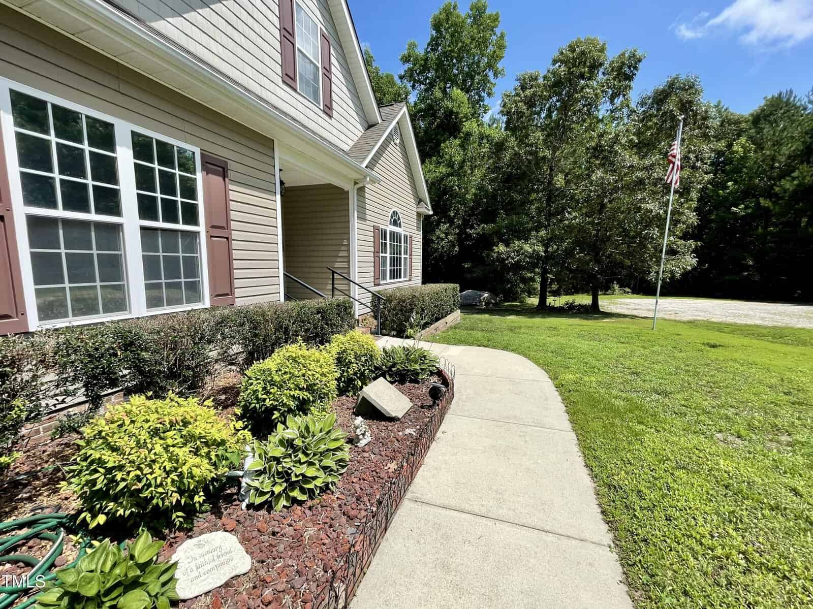 A concrete walkway curves past landscaped shrubs and plants toward the entrance of a tan house with white trim. An American flag stands on a pole on the grassy lawn, surrounded by trees under a blue sky.