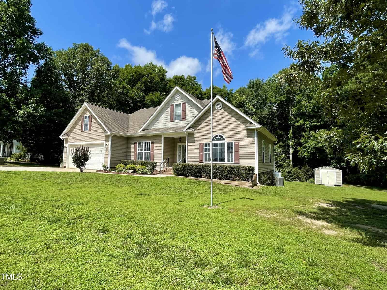 Single-story beige house with gabled roof, white trim, and front porch, set on a large green lawn. An American flag is on a pole in the yard, with trees and a small shed in the background under a blue sky.