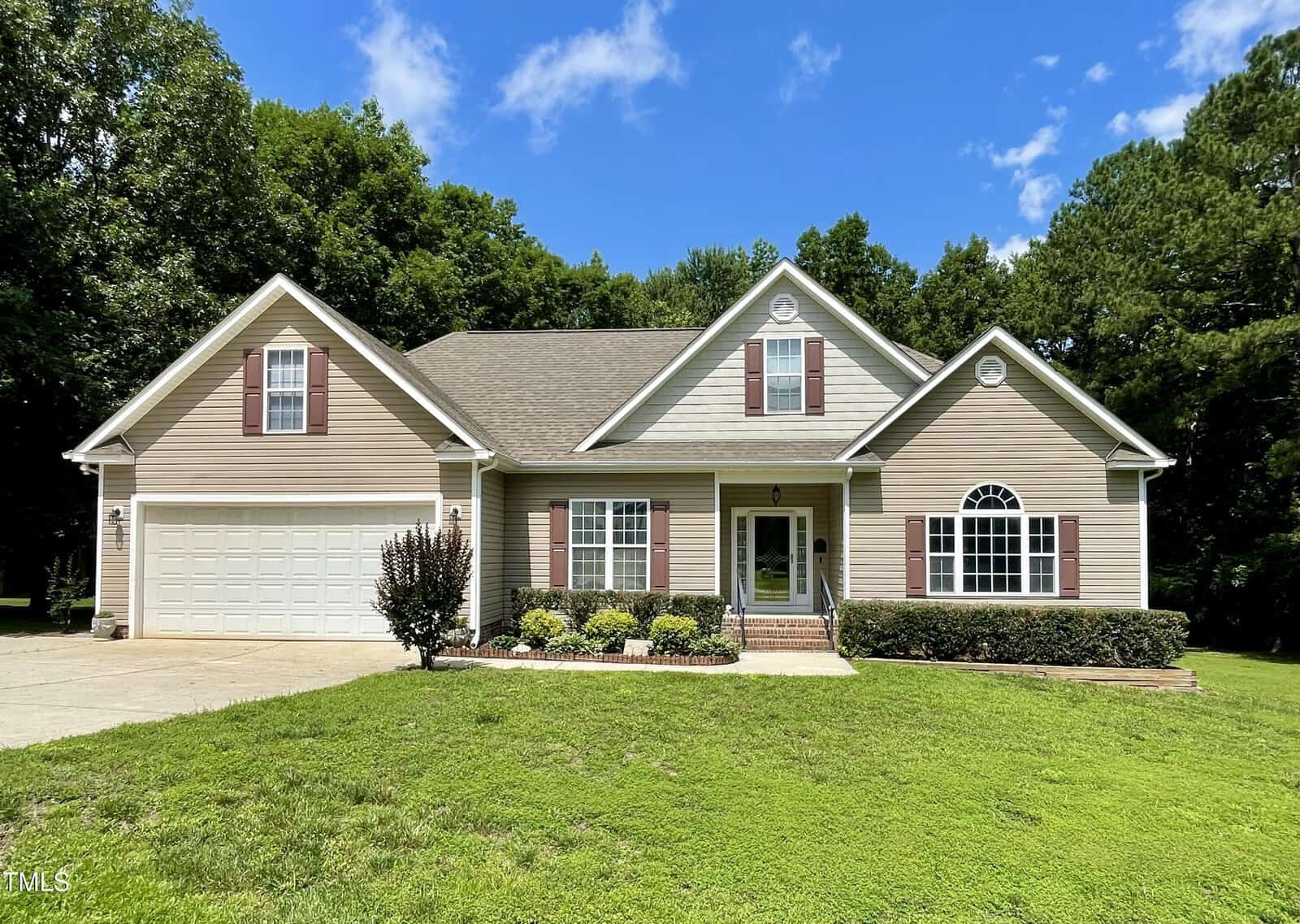 Single-story beige house with white trim, brown shutters, a large arched window, and a two-car garage, set on a spacious green lawn with trees and blue sky in the background.