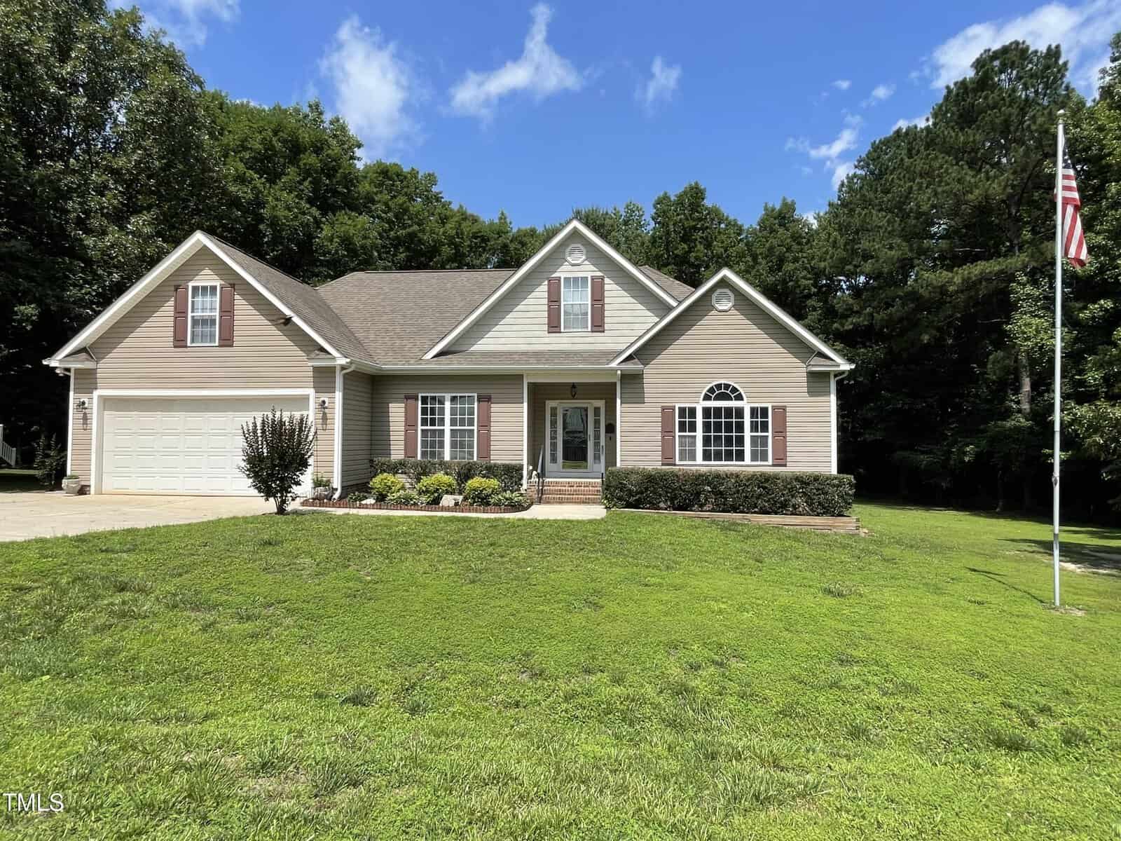 Single-story tan house with white trim and a gabled roof, set on a large green lawn. There is a garden with shrubs in front, a driveway to the left, and an American flag on a pole to the right. Trees border the background.