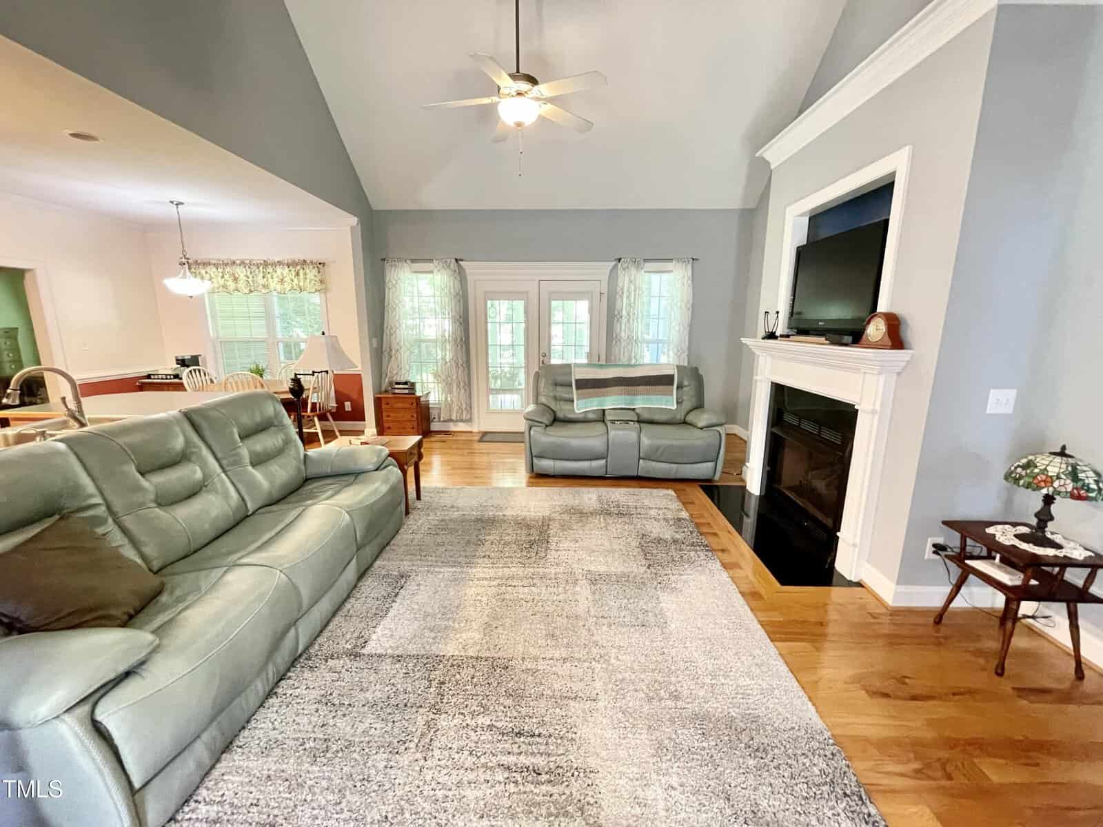 Living room with gray sofas, large area rug, hardwood floors, a white fireplace with TV above, ceiling fan, and French doors with curtains leading outside. Dining area and kitchen are visible to the left.