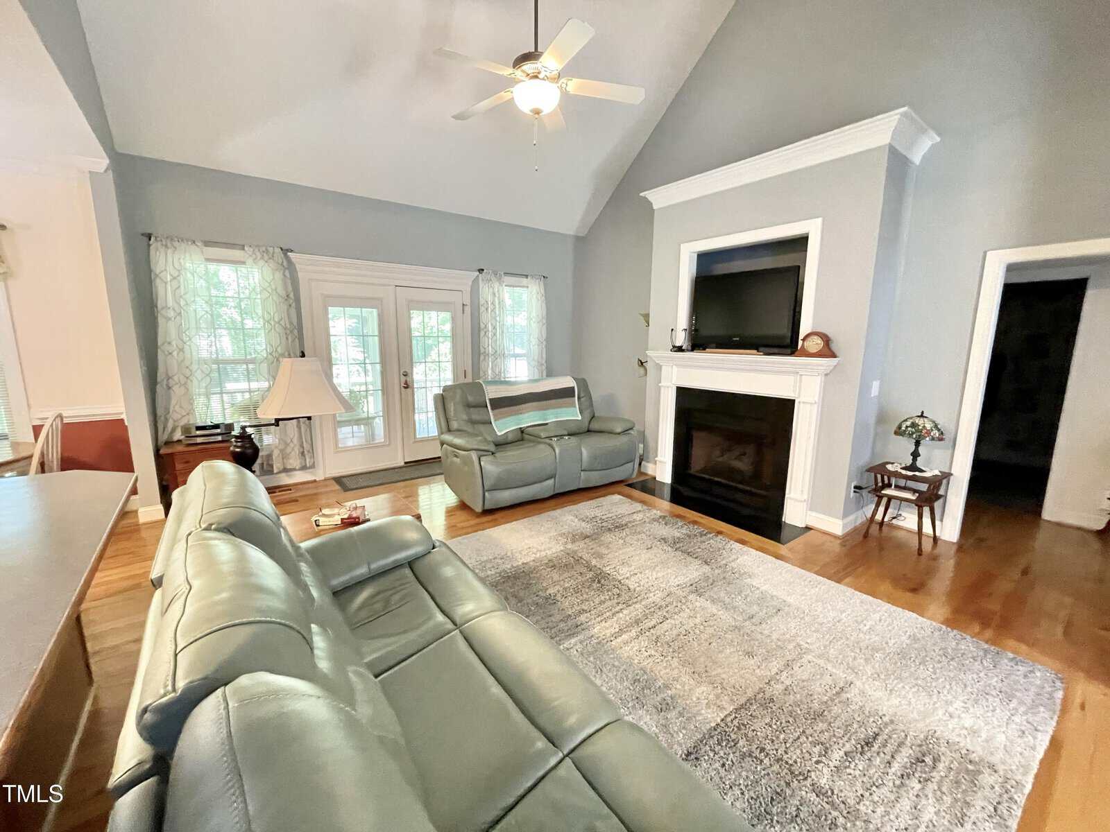 Spacious living room with gray sofas, a large area rug, fireplace with a TV above, hardwood flooring, and windows with sheer curtains. Ceiling fan is centered above the room, which features high, sloped ceilings.