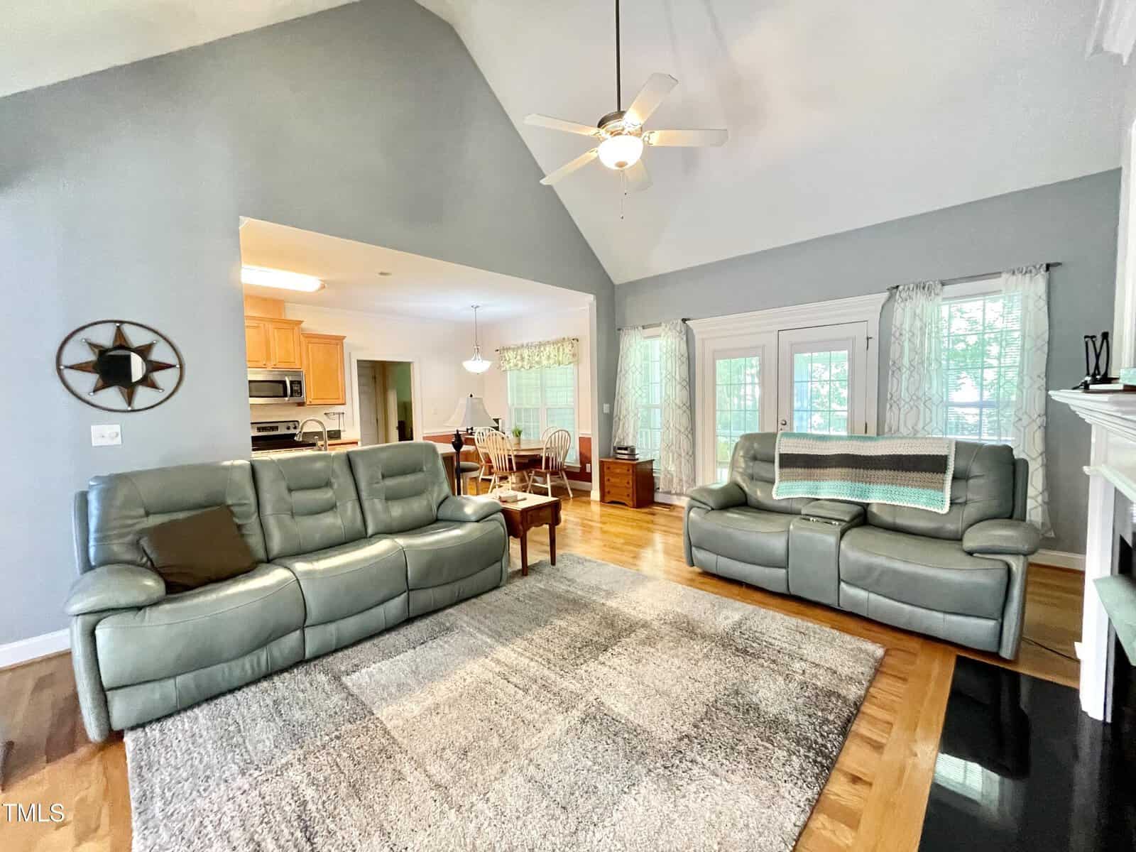 A living room with two gray leather sofas, a ceiling fan, a gray area rug, and hardwood floors. The space opens to a kitchen and dining area with wooden cabinets and a dining table. Light streams in through multiple windows.