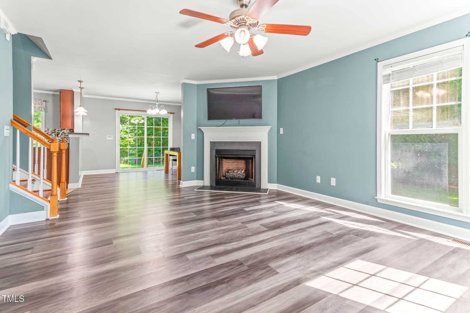 A spacious living room with light gray wood flooring, teal walls, a white-framed window, ceiling fan, and a fireplace below a mounted TV. Dining area and kitchen with large windows are visible in the background.