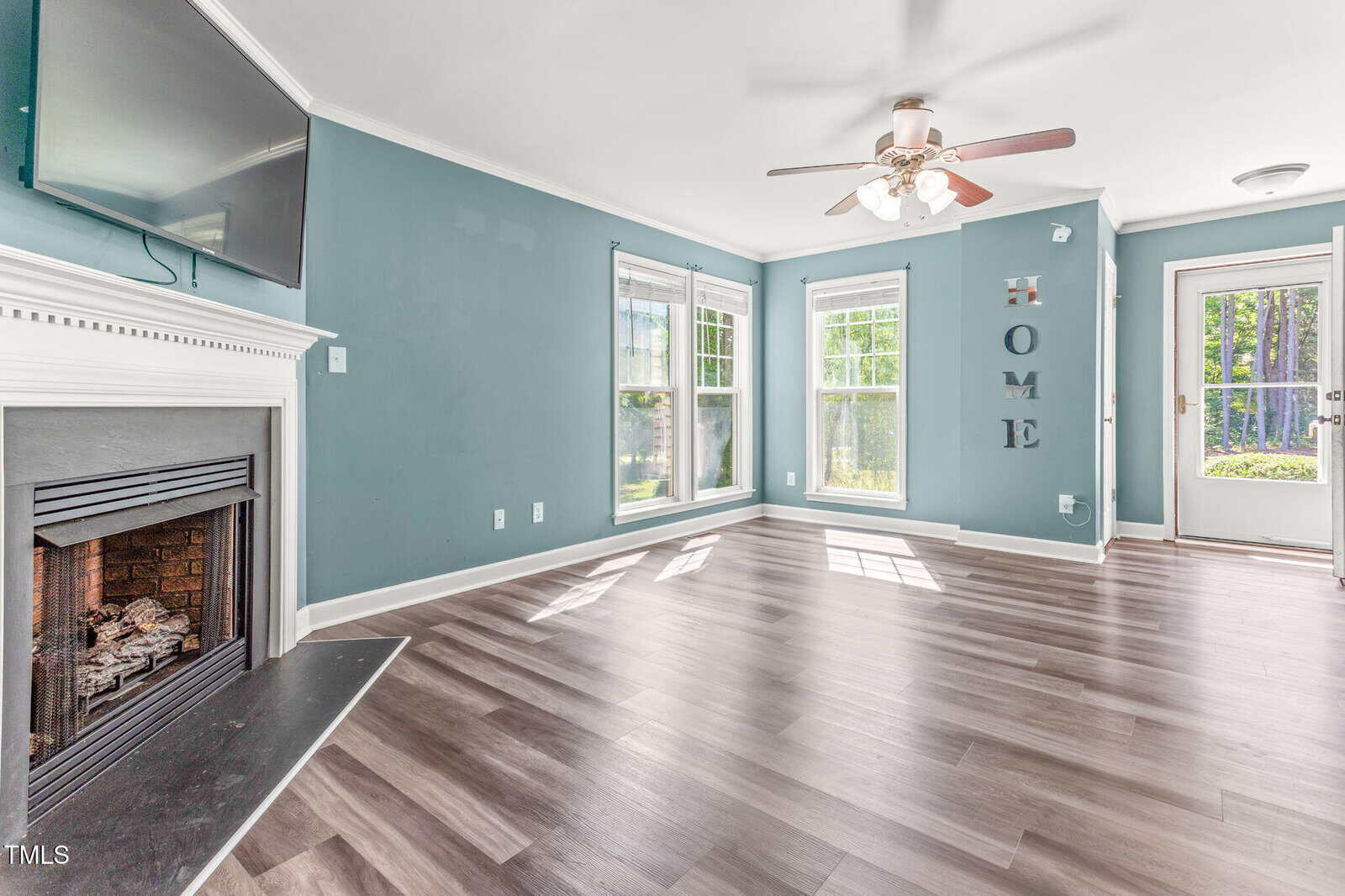 A bright living room with blue walls, wood-look flooring, a fireplace with a TV above it, three large windows, a ceiling fan, and a door leading outside. The word "HOME" is mounted on the wall.