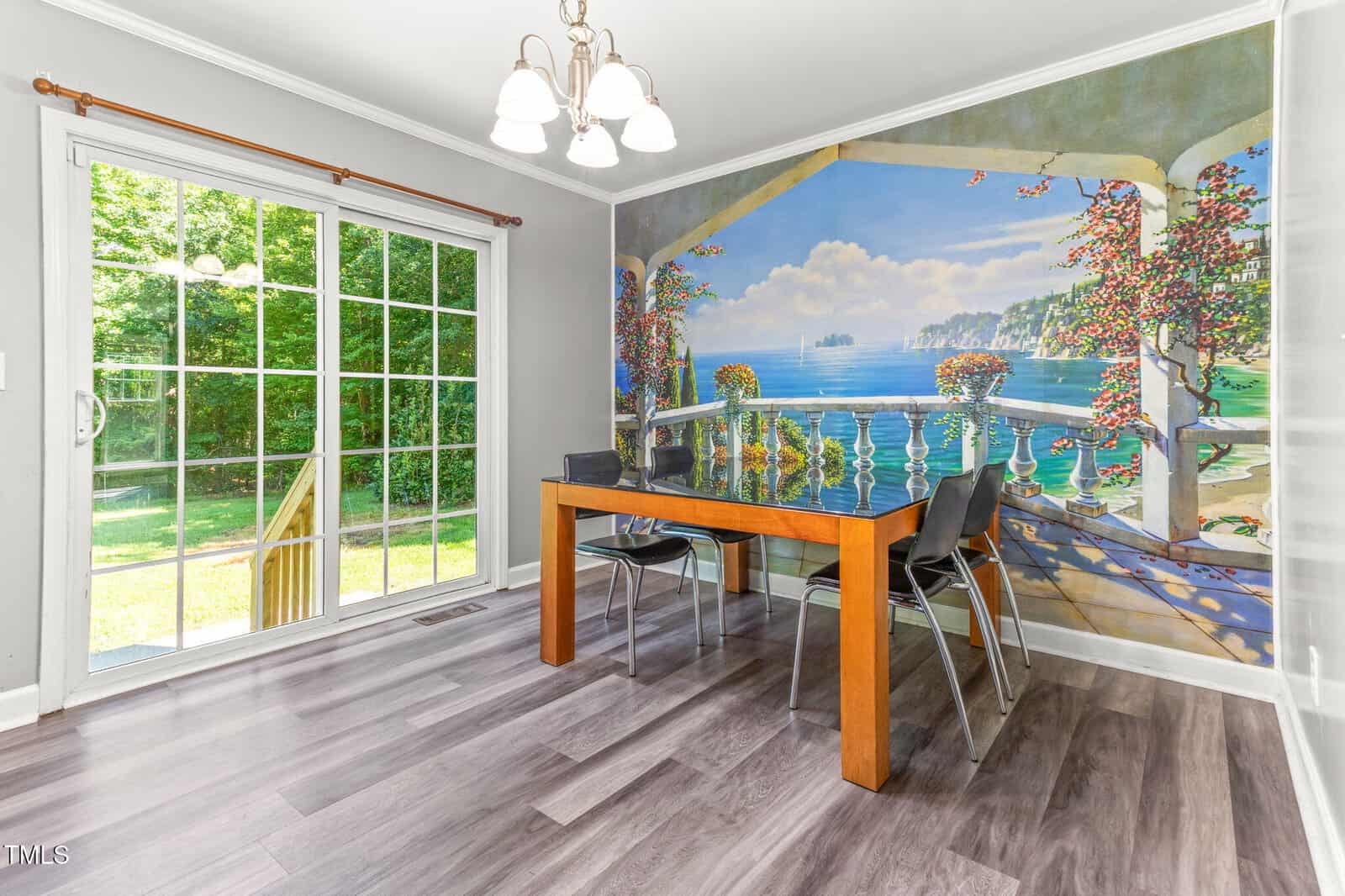 A dining area with a rectangular wooden table and four black chairs sits on gray wood-look flooring. A mural of a seaside balcony scene covers one wall, and large glass doors open to a green yard outside.