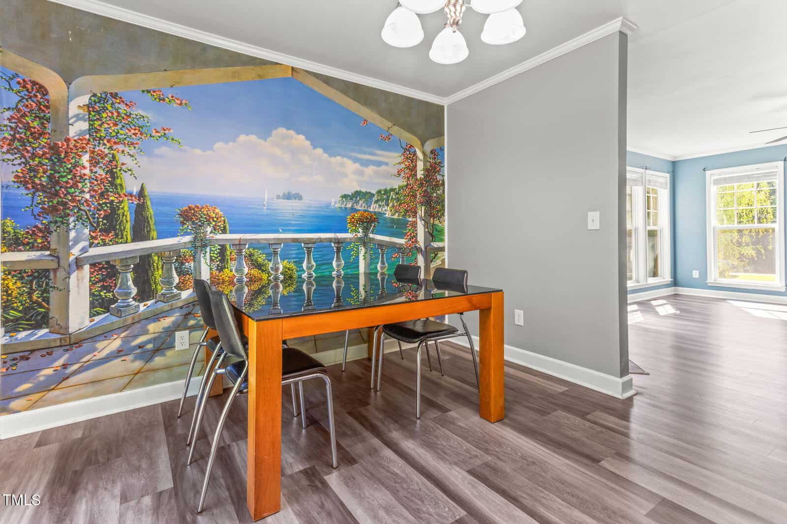 A small dining area with a wooden table and four black chairs is set against a large wall mural depicting a scenic ocean view from a terrace. The room has gray walls and light-colored wood flooring.