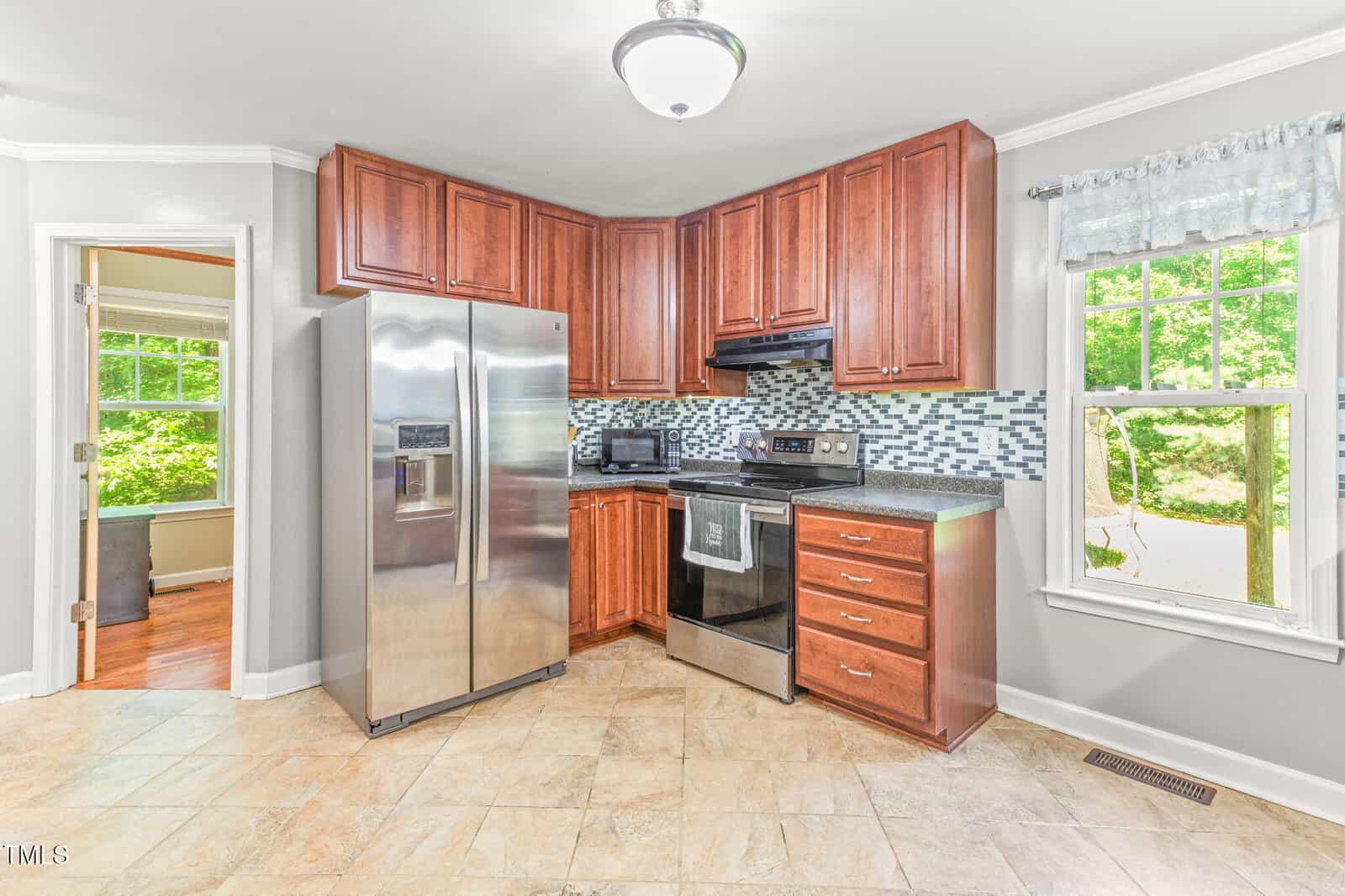 Modern kitchen with stainless steel refrigerator, oven, and microwave, brown cabinets, tile floor, and a window with a view of green trees. Doorway leads to another room with wood flooring.