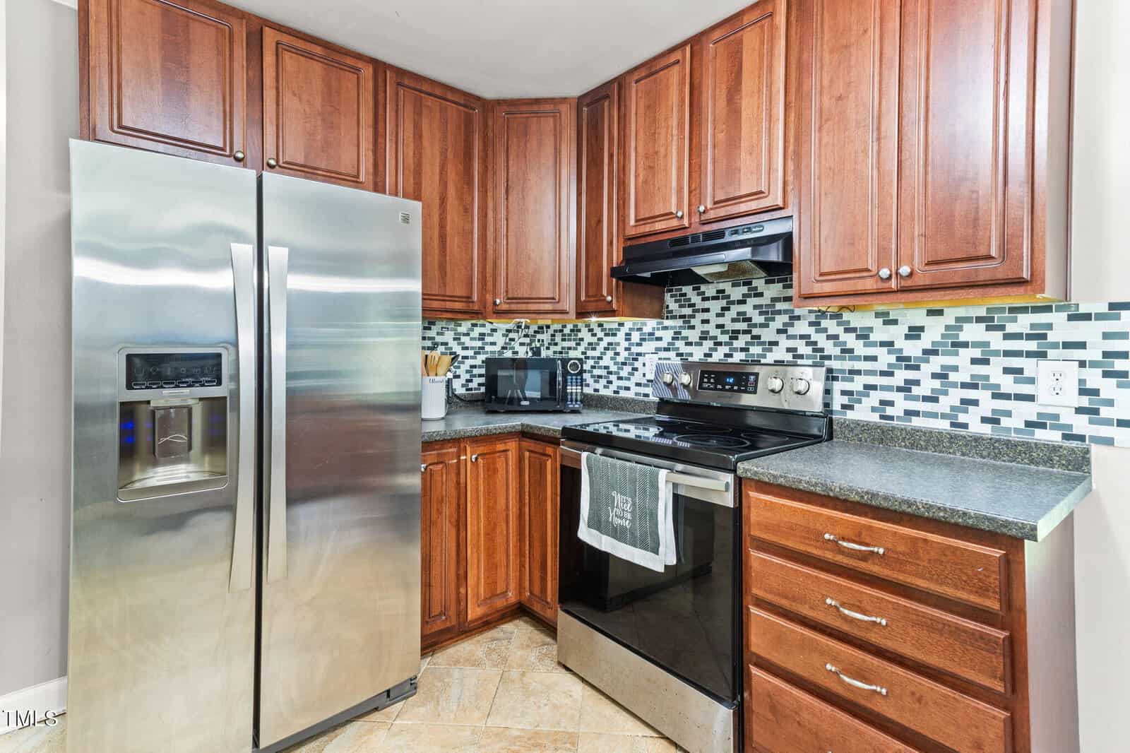 Modern kitchen with stainless steel refrigerator, electric stove, black range hood, microwave, and wooden cabinets. The backsplash features a mosaic tile pattern in shades of gray, white, and blue.