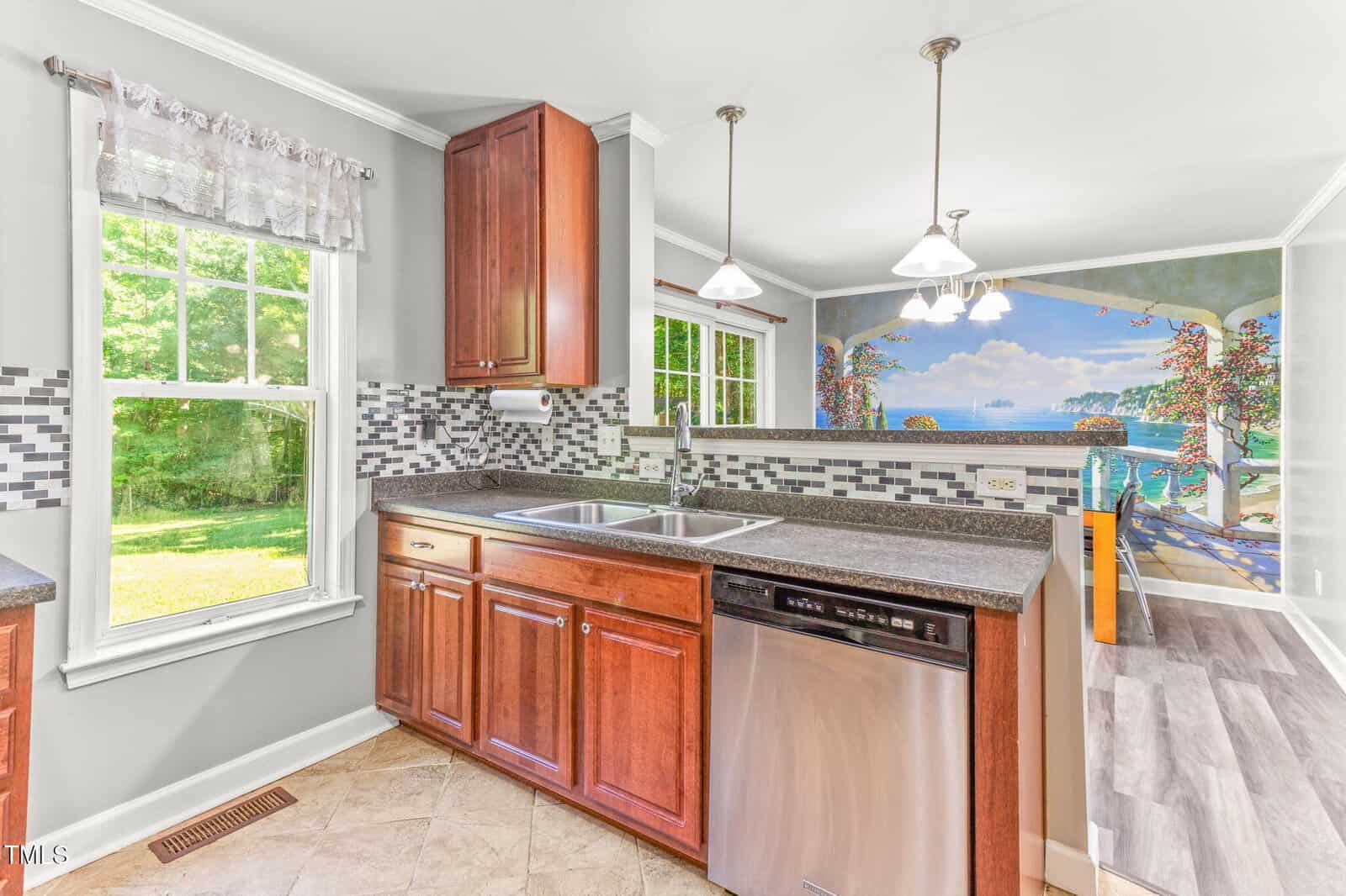 A kitchen with brown cabinets, a double sink, and stainless steel dishwasher. A window allows sunlight in. The adjacent room features a mural of a beachside scene with trees, water, and a blue sky.
