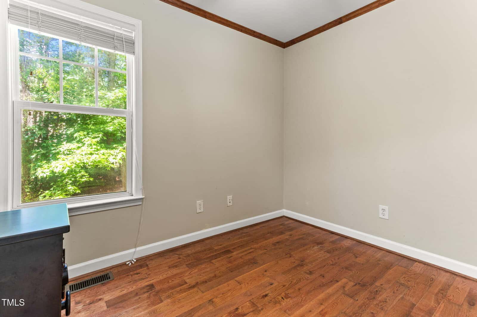 Empty room with light beige walls, wooden floor, a window with blinds letting in sunlight, white trim, and a small black dresser partially visible in the bottom left corner.