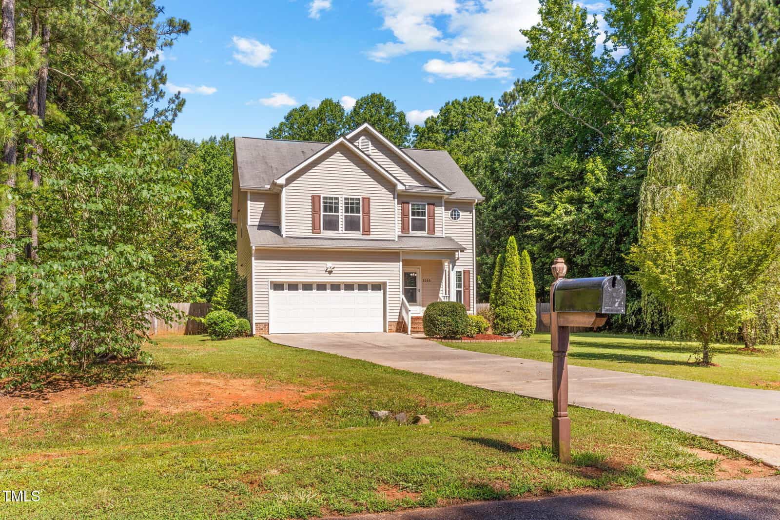 A two-story beige house with white trim and a double garage sits at the end of a concrete driveway, surrounded by trees and a lawn. A mailbox is positioned at the edge of the driveway near the street.