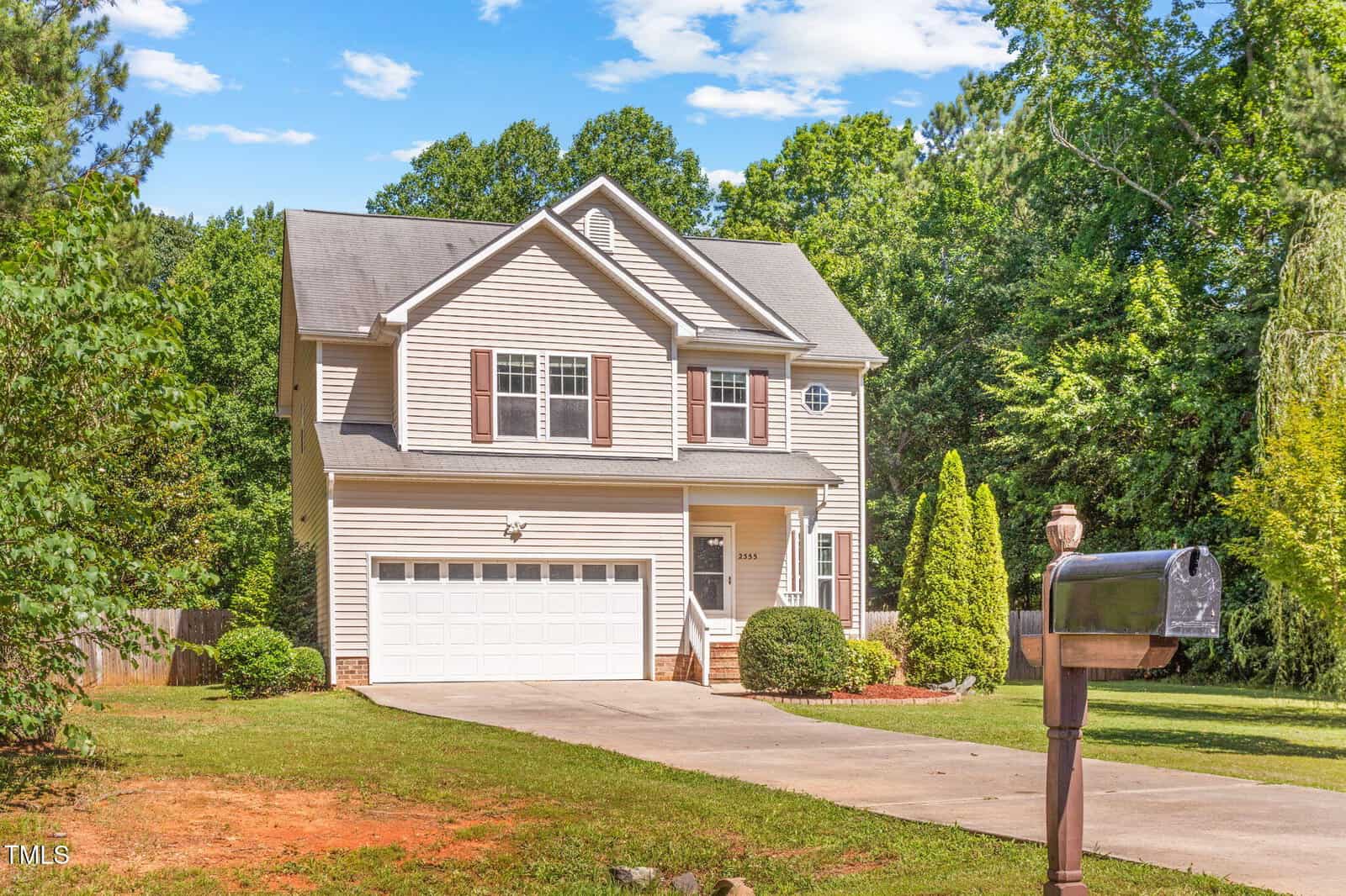 A two-story beige house with brown shutters and a white garage door, surrounded by trees and greenery. A black mailbox is visible in the foreground near the driveway.