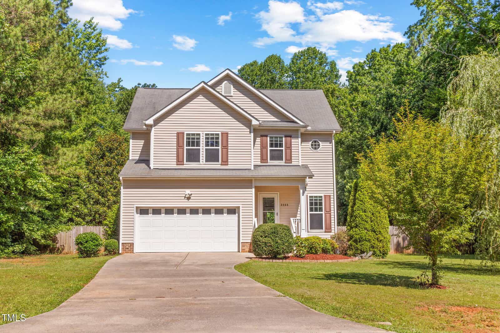 Charming two-story beige house with white trim at 2555 Winding Creek Lane, Franklinton. Features a two-car garage, concrete driveway, lush front yard with shrubs and small trees, and wooded areas in the background under a partly cloudy sky.
