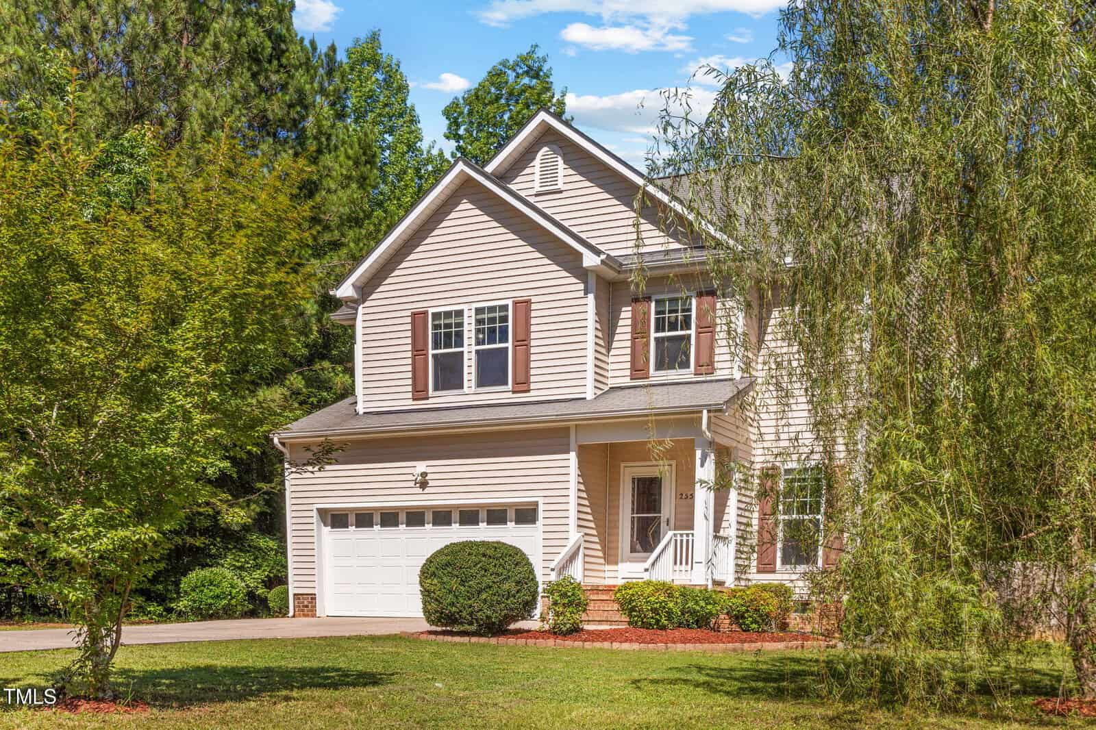 Two-story beige house with white trim, brown shutters, and a front porch. The house has a two-car garage and is surrounded by green trees and grass under a blue sky.