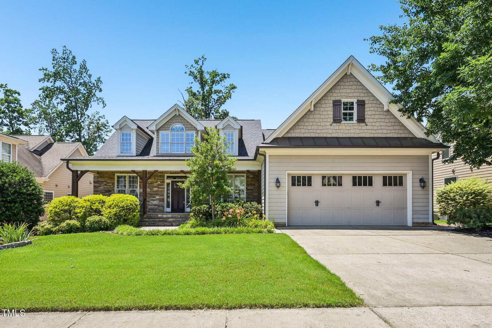 A suburban house with beige siding, stone accents, dormer windows, a manicured lawn, shrubs, and a double garage, set under a clear blue sky.