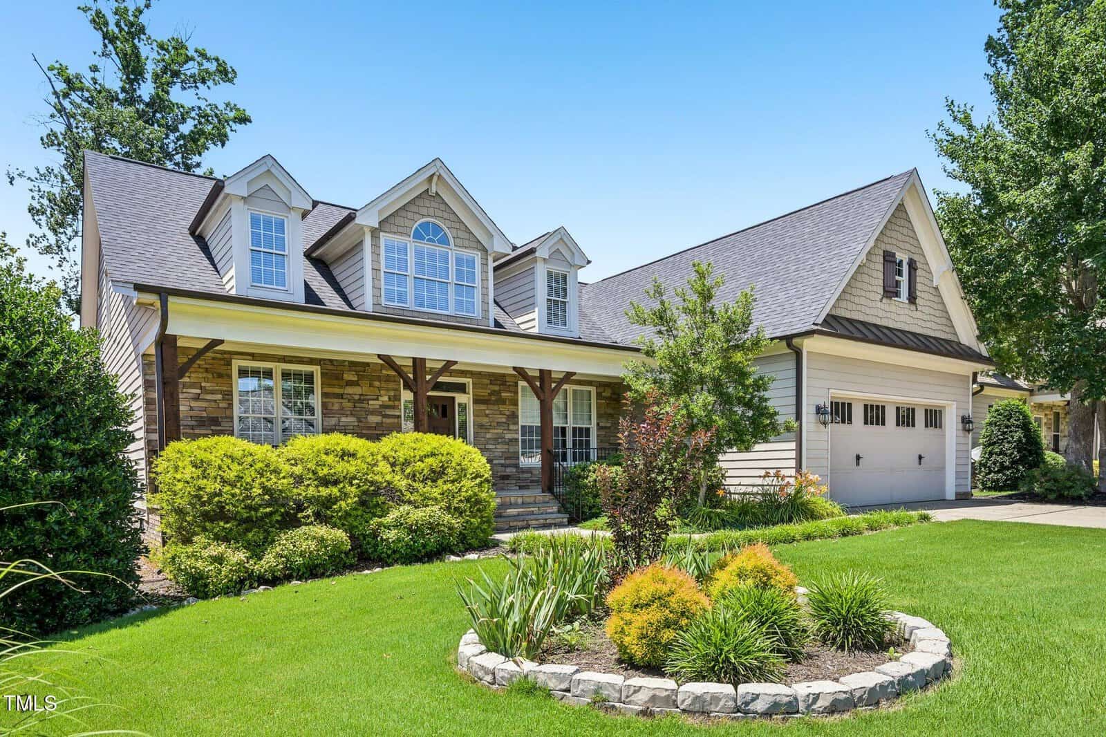 A two-story suburban house with a front porch, stone and siding exterior, manicured lawn, landscaped garden beds, and a double garage under a clear blue sky.