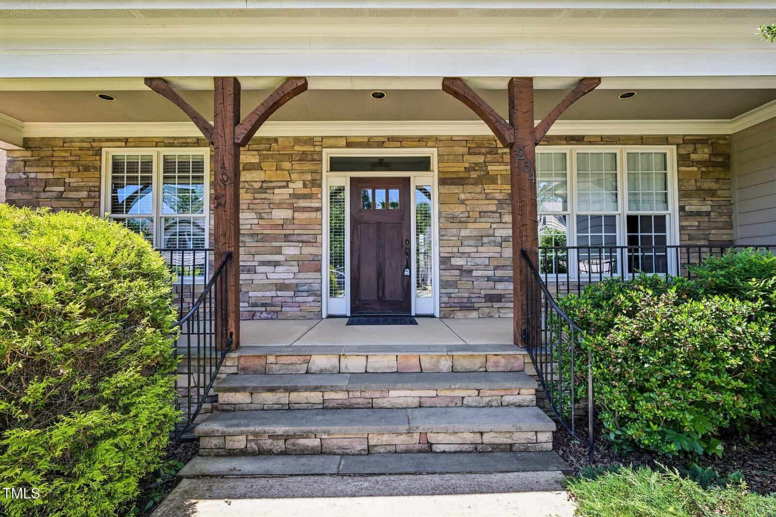 A front porch with stone steps, metal railings, a wooden door, and large windows on either side. The house exterior features stonework and neatly trimmed bushes flank the entrance.