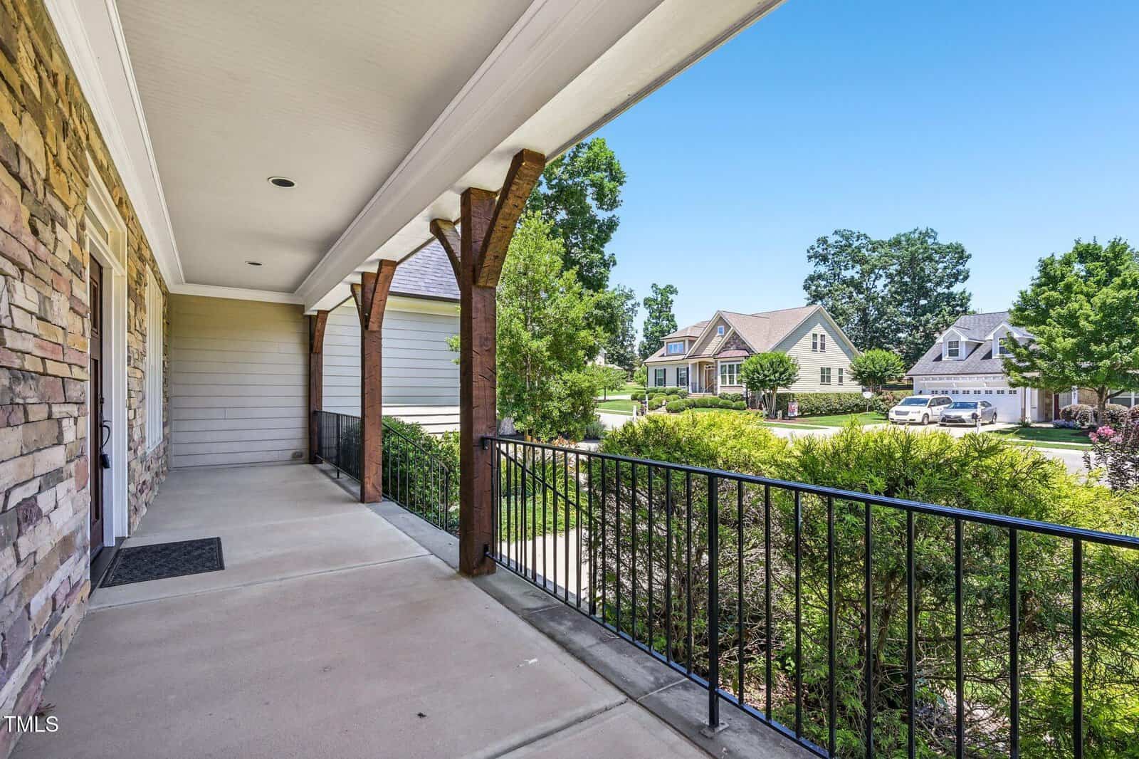 View from a covered front porch with stone and wood accents, metal railing, and a concrete floor, overlooking a suburban street with houses, green trees, and a clear blue sky.