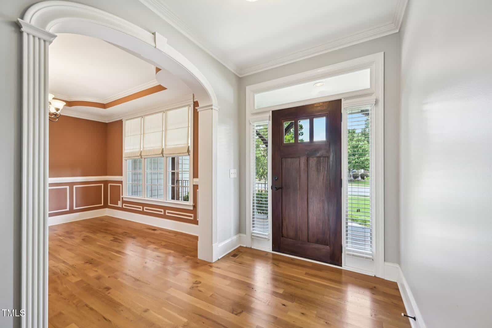 A bright foyer with a dark wooden front door, sidelights, and transom window, opens to a dining room featuring large windows, brown and white wainscoting, hardwood floors, and crown molding.