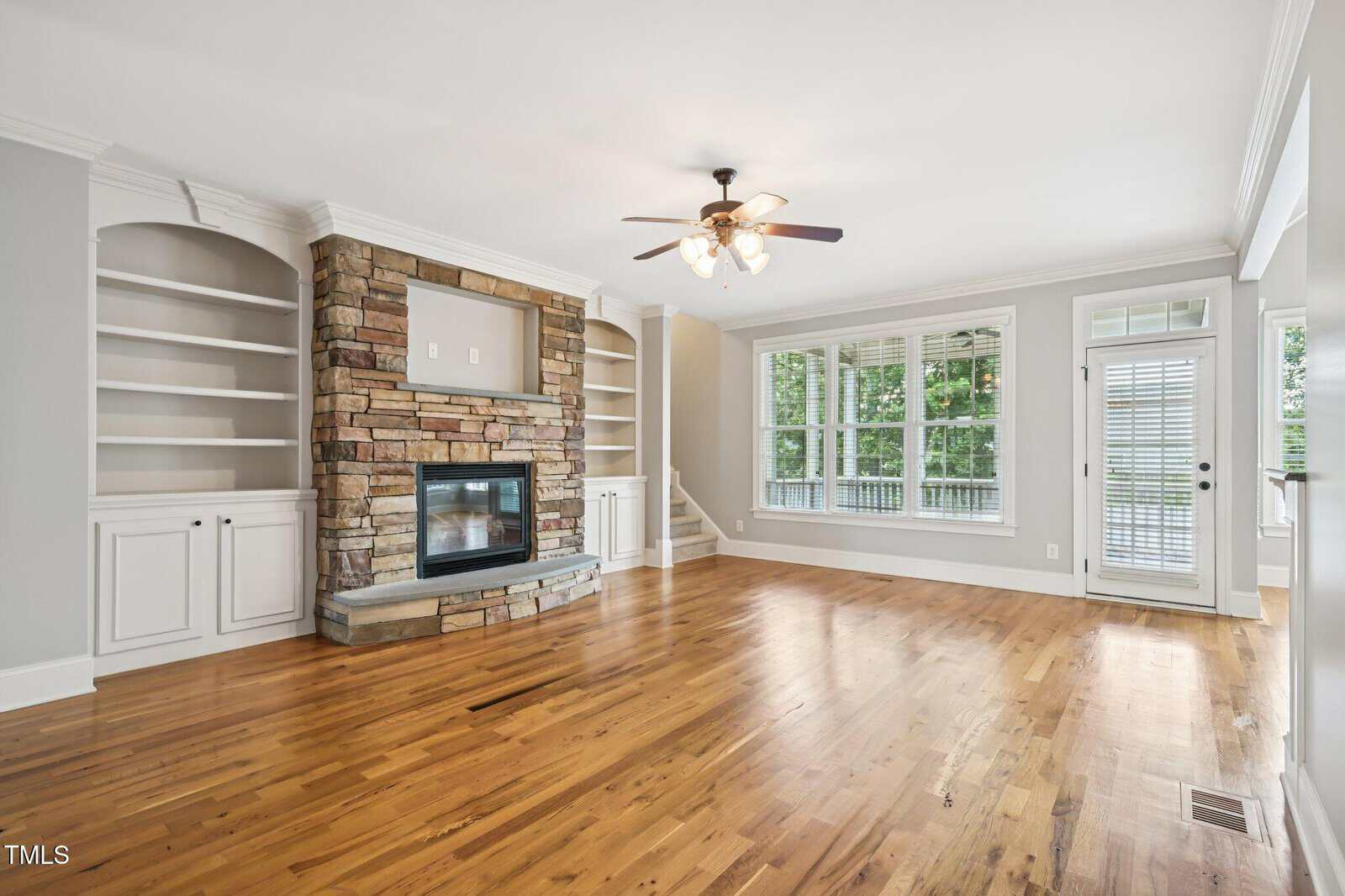 Bright living room with hardwood floors, a stone fireplace flanked by built-in shelves and cabinets, large windows, a ceiling fan with lights, and a glass door leading outside. Walls are light gray with white trim.