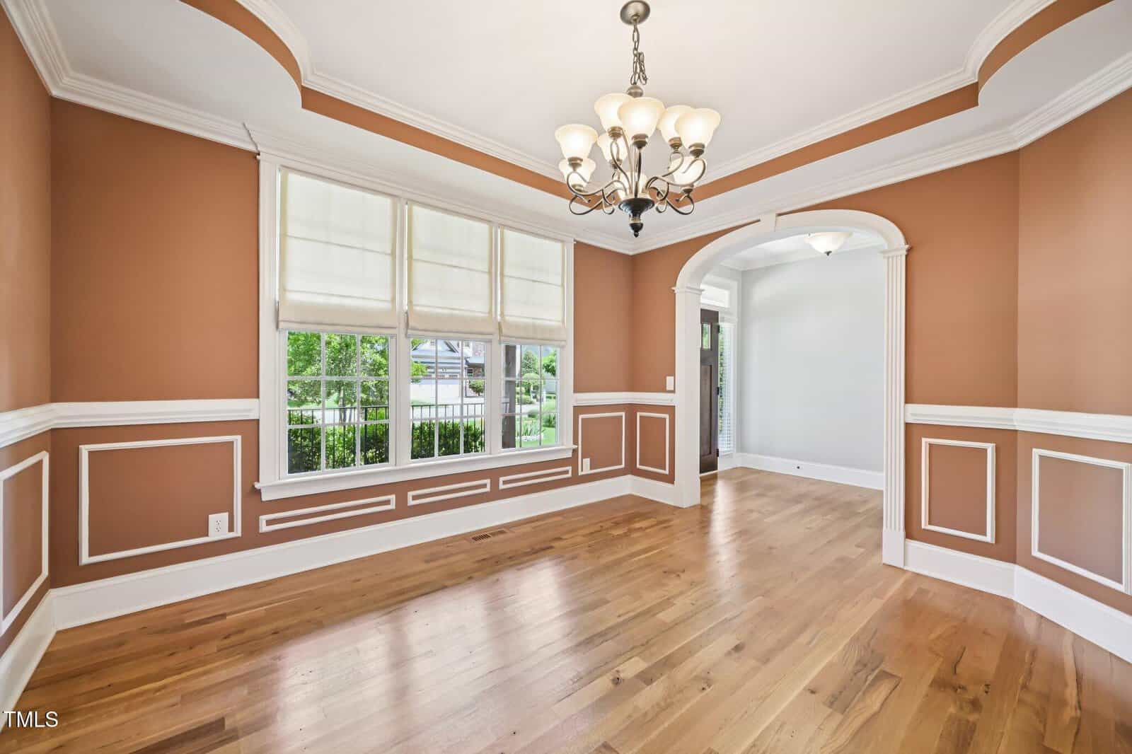 A spacious dining room with hardwood floors, peach-colored walls with white trim, large windows with shades, a chandelier, and an arched entryway leading to another room.
