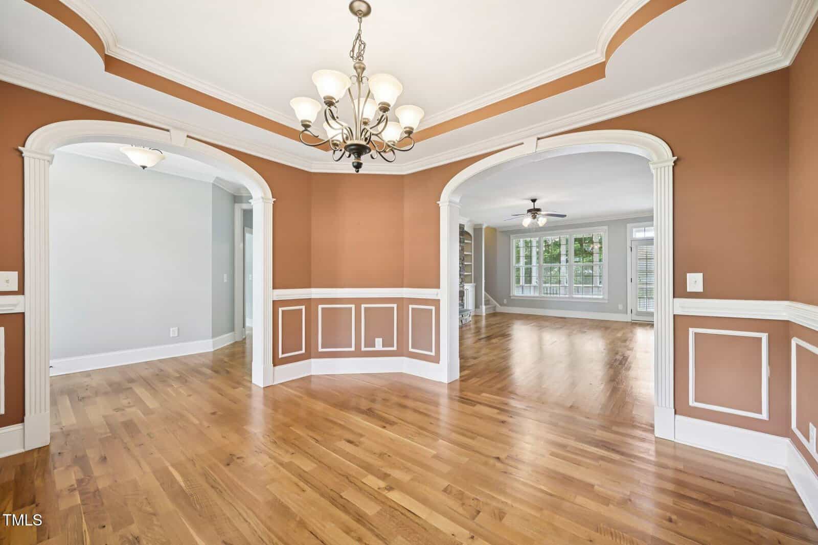 Dining room with hardwood floors, white trim, two archways, tan and white walls, white ceiling with crown molding, and a chandelier. The room opens to a hallway and a living space with large windows.