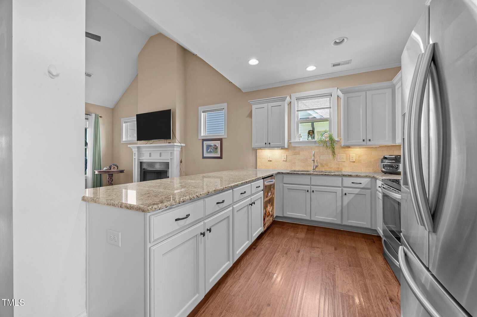 A modern kitchen with white cabinets, stainless steel appliances, granite countertops, and wood flooring. The kitchen island faces a living area with a fireplace and a wall-mounted TV. Natural light enters through a window above the sink.
