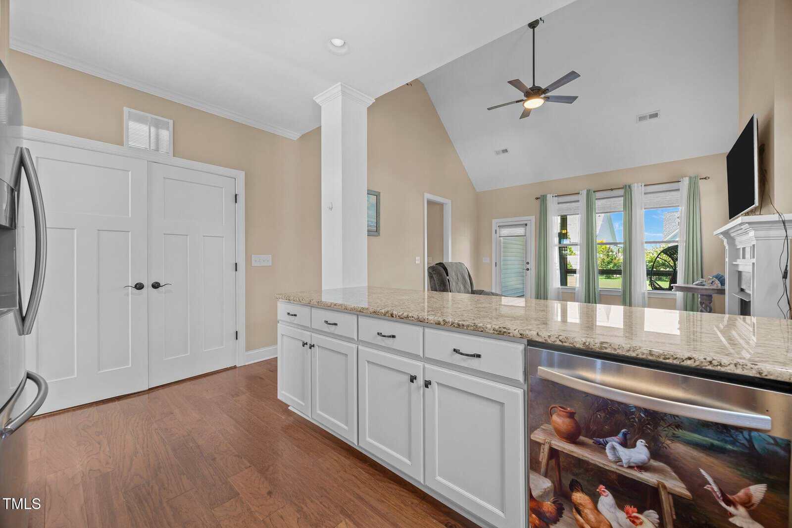 A modern kitchen with white cabinets, a granite island countertop, hardwood floors, a double door pantry, and a refrigerator decorated with a painted farm animal scene. Ceiling fan and large windows are visible in the background.