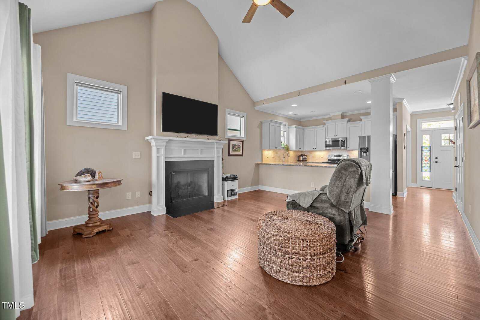 Open living space with hardwood floors, a fireplace under a wall-mounted TV, a small round table, a recliner, and a wicker ottoman. The kitchen with white cabinets is visible in the background. Natural light fills the room.