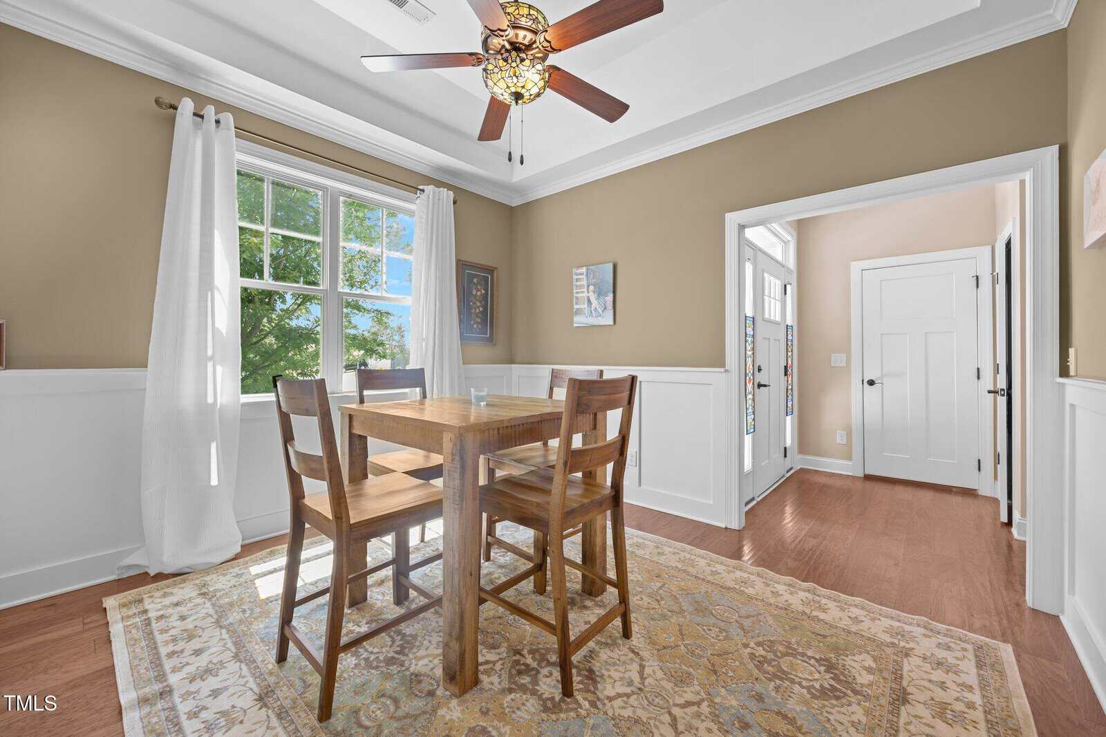 A small dining room with a wooden table and four chairs on a patterned rug, tan walls with white trim, a window with white curtains, ceiling fan, and artwork on the walls. Hardwood floor and view of an adjacent hallway.