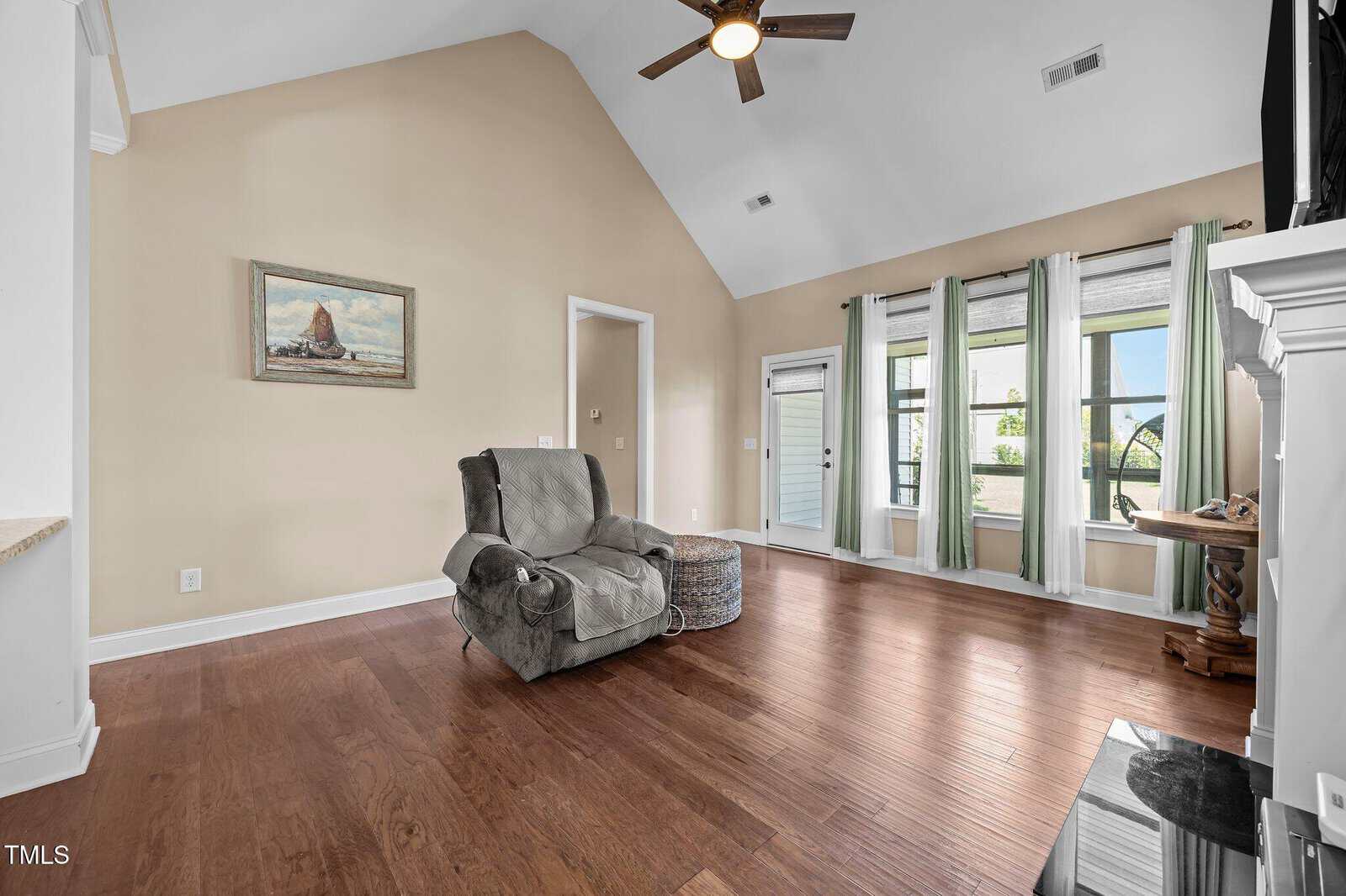 A spacious living room with vaulted ceiling, hardwood floors, a recliner chair, a woven ottoman, and large windows with white curtains letting in natural light. A ceiling fan is mounted above.