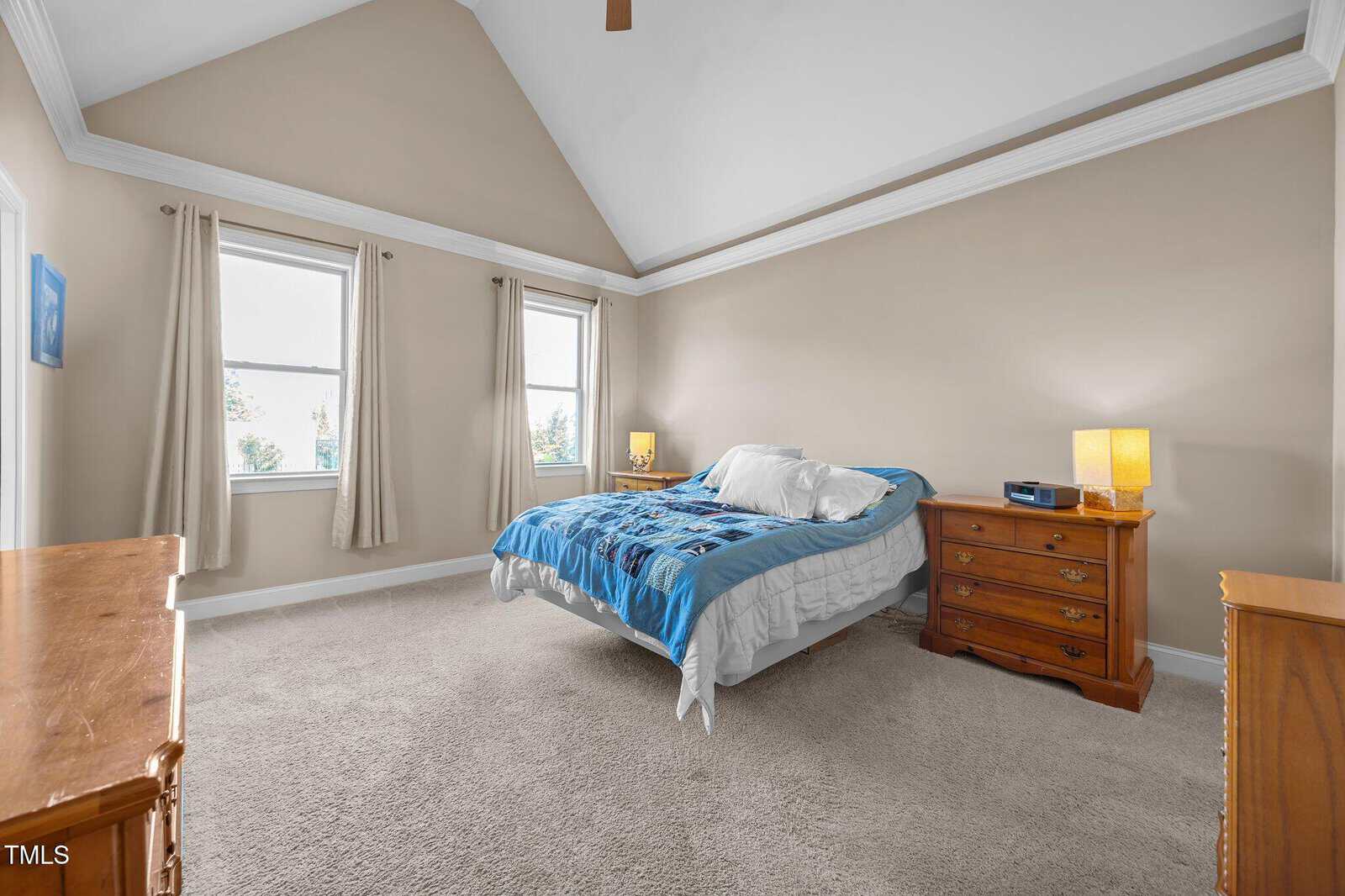 A bedroom with beige walls, a vaulted ceiling, carpeted floor, a bed with blue bedding, two nightstands with lamps, and a dresser. Two windows with curtains let in natural light.