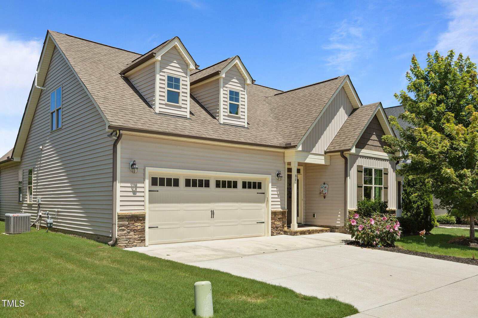 A modern suburban house with beige siding, stone accents, two dormer windows, a double garage, and a well-maintained lawn with shrubs and a blooming bush by the front entrance.