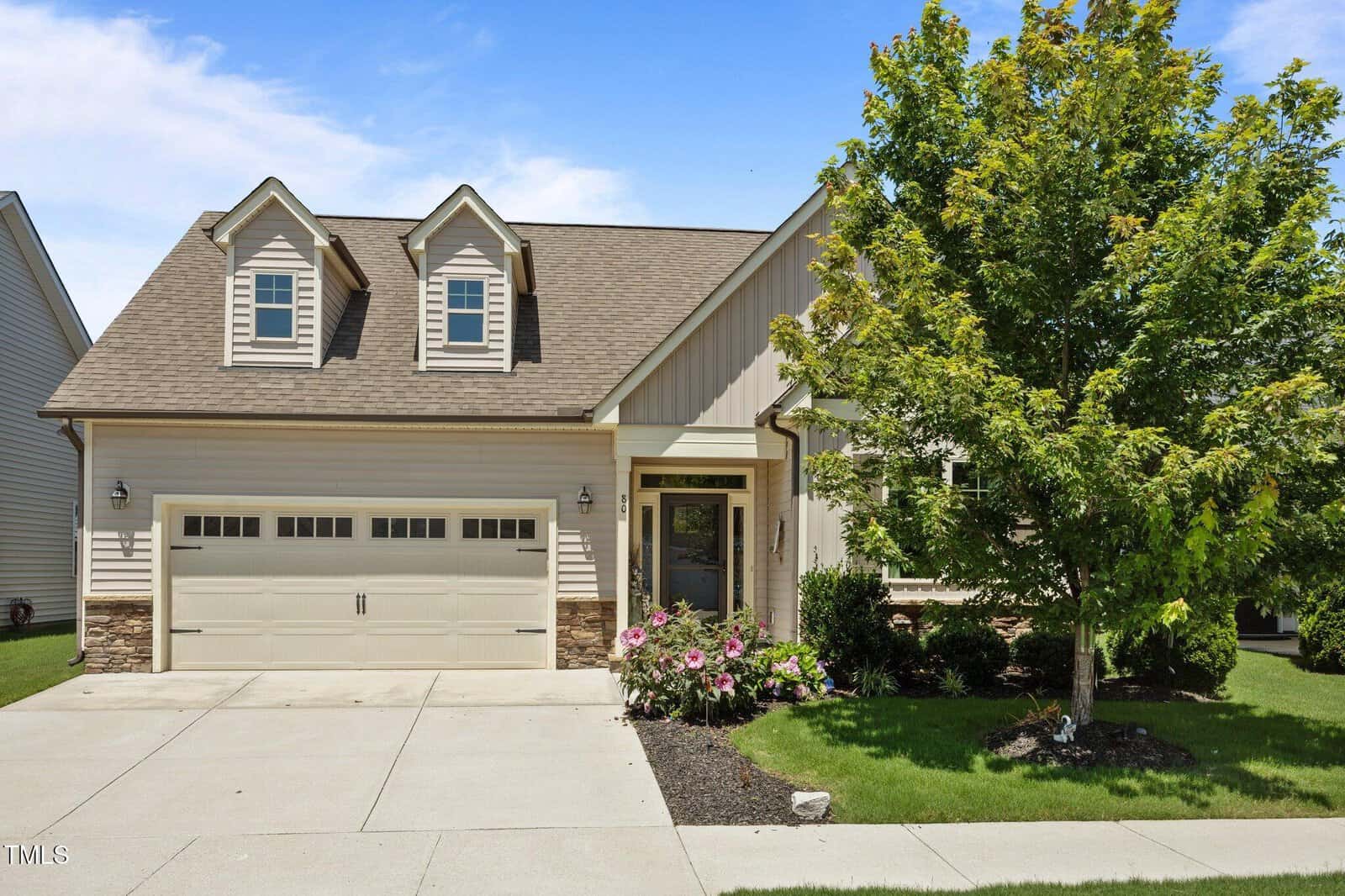 A modern suburban house with beige siding, two dormer windows, a double garage, manicured lawn, flowering shrubs, and a large green tree, all under a blue sky with light clouds.