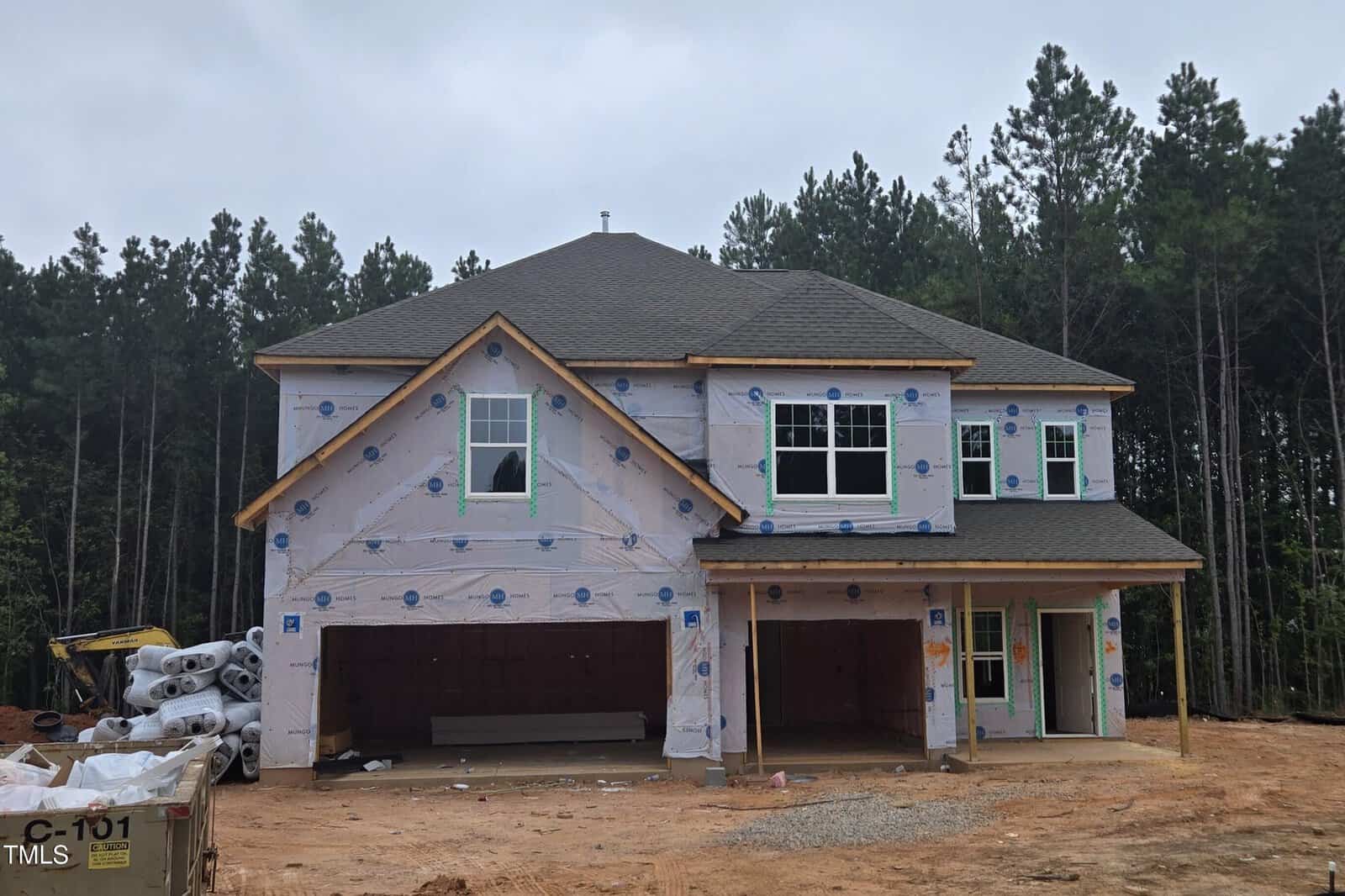 Two-story house under construction, wrapped in weatherproofing material with exposed wooden beams. Windows and garage doors are installed. Surrounded by dirt and construction materials, with trees in the background.