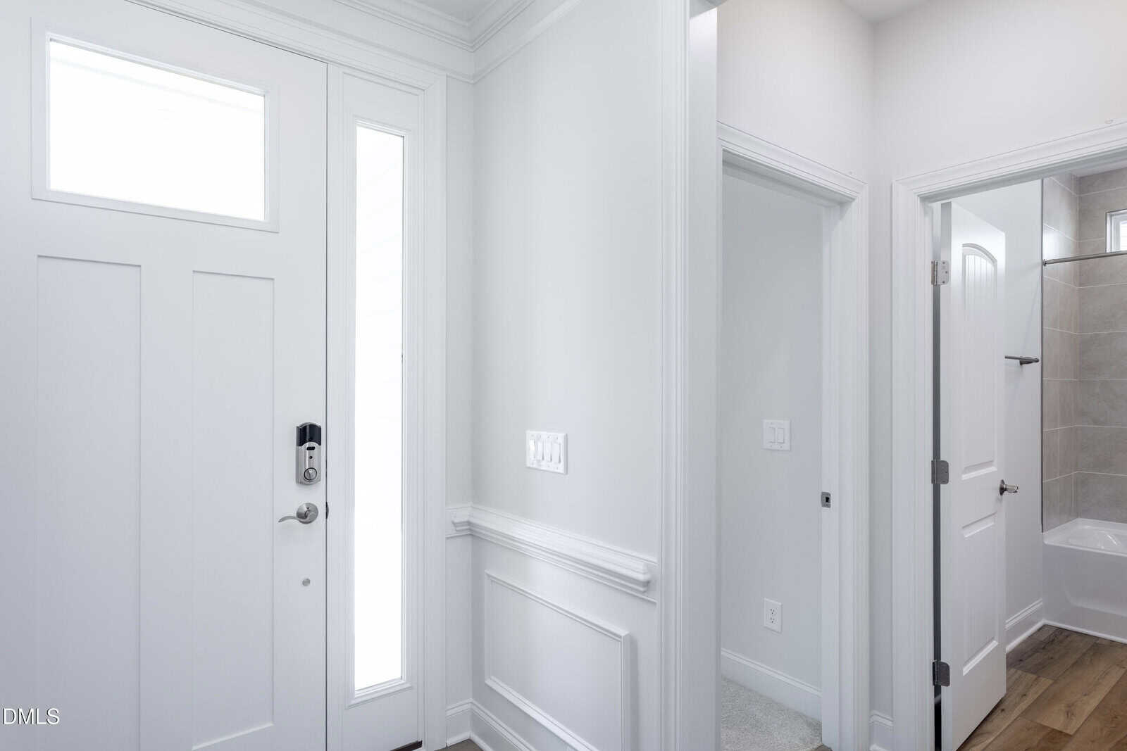 A bright, modern entryway with a white front door featuring a narrow vertical window, paneled walls, hardwood floors, and a hallway leading to a bathroom with light tile and a visible bathtub.